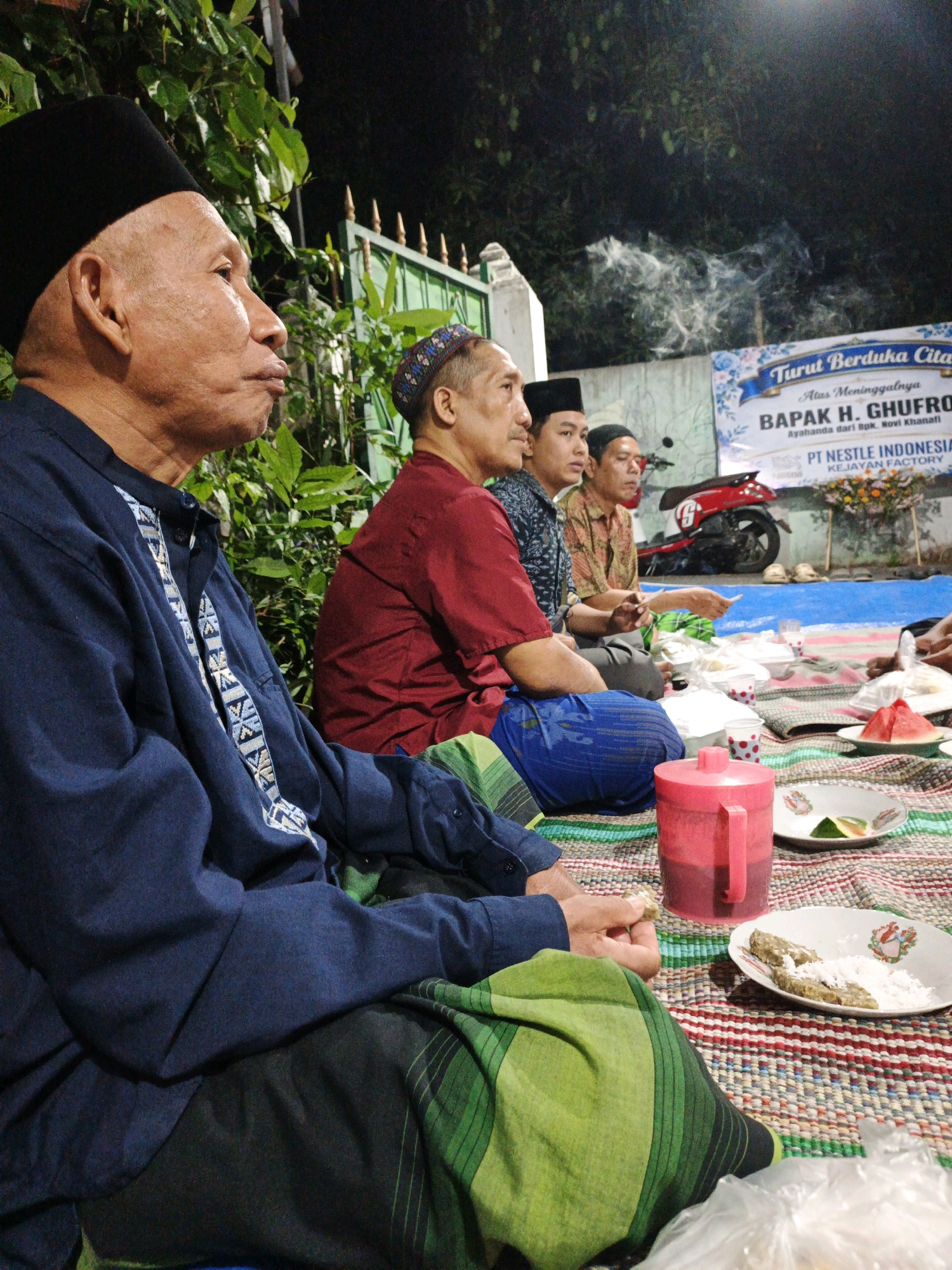 The image shows a gathering of men sitting on a mat outdoors at night. There are trees and a fence in the background, and a banner that reads "Turut Berduka Cita" (Condolences) with text below it that appears to be in Indonesian. In the foreground, there are plates of food and a red pitcher. Several men are wearing traditional Indonesian attire. A red motorcycle is parked behind them. The scene appears to be a memorial or prayer gathering.