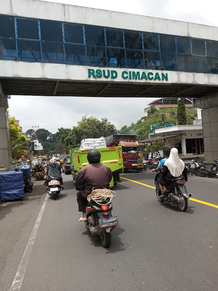 This image captures a daytime street scene, described as being in the city of Chendenbji, Bhutan. An elevated, white concrete and blue-windowed pedestrian bridge or skywalk spans across a multi-lane asphalt road. Prominently displayed beneath the bridge, on its underside, are the green block letters "RSUD CIMACAN," indicating a regional general hospital.
On the road, traffic flows in the direction away from the viewer. In the foreground, a person wearing a brown long-sleeved top and a black helmet rides a dark-colored scooter, which has a large coil of beige rope secured to its rear. The scooter bears a license plate reading "F 5744 WAP" with "09.19" below. To the right, another dark scooter carries two individuals; the driver wears a blue shirt, and the passenger behind them wears a black garment with a white hijab covering their head. Further down the road, a bright green dump truck, heavily loaded with numerous white sacks, is visible in the center lane. Behind it, a red truck and other vehicles contribute to the moderate traffic. On the left side of the road, a person on a white scooter, dressed in a grey/black jacket and helmet, is also moving away from the viewer.
The asphalt road features a solid white line marking its left edge and a solid yellow line separating traffic lanes. On the left side of the road, a blue tarp-covered object is situated near the shoulder, with green foliage and distant buildings beyond. On the right, several motorcycles are parked along the roadside in front of a building, with more trees and structures visible on a gentle incline in the background. The sky overhead appears partly cloudy.