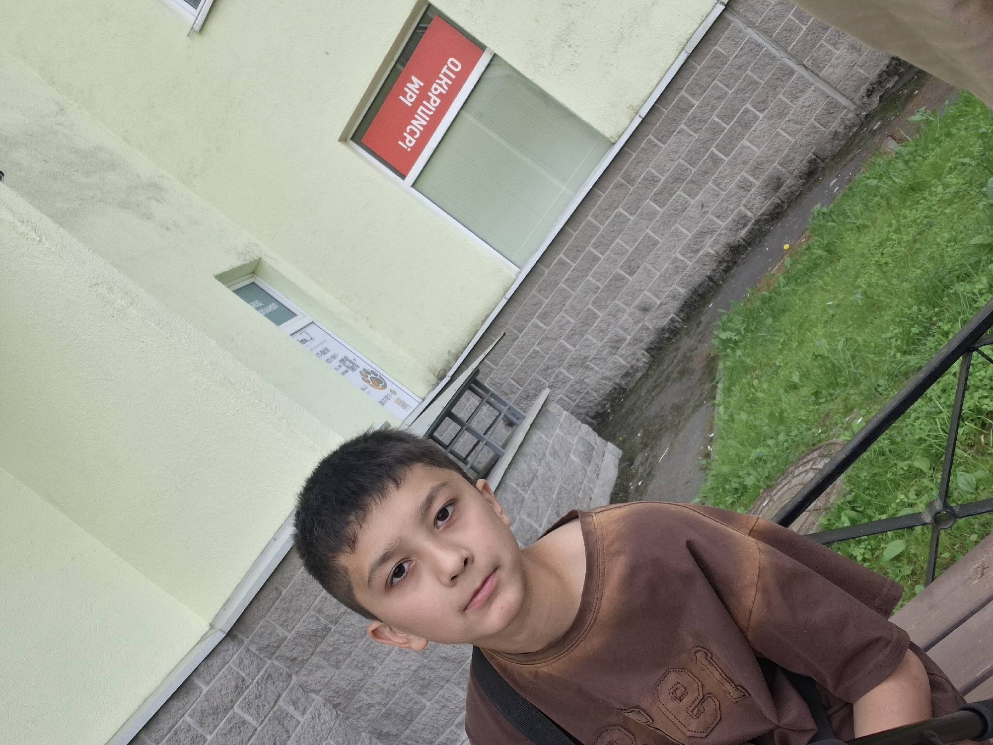 The image features a young boy, appearing to be in his early teens, looking directly at the camera with a neutral expression. He has short dark hair, dark eyes, and is wearing a plain brown t-shirt with some subtle textured lettering or design, part of which reads "EL" or "LE". A black strap, likely from a bag, is visible over his shoulder.
The setting is outdoors, next to a building in Saint Petersburg, Russia. The building's upper facade is covered in a light greenish-yellow textured stucco, while the lower part is clad with gray rectangular stone or brick tiles. Two windows are visible on the building. The upper window displays a red sign with white text, which, when properly oriented, reads "ОТКРЫТО!" (OPEN!) and "WPI". The lower window also contains faint white text, identifiable as "СУВЕНИРЫ" (SOUVENIRS) and "ПАРФЮМЕРИЯ" (PERFUMERY), along with other smaller details like numbers.
To the boy's right, a dark brown wooden bench with black metal railings is partially visible. The ground consists of a paved area with gray rectangular stones next to the building, leading to a patch of green grass. Some areas within the grass appear muddy or dirty, and a small yellow flower can be seen. The lighting suggests an overcast day. The photograph is taken from a slightly elevated vantage point, looking down towards the boy and the immediate surroundings.