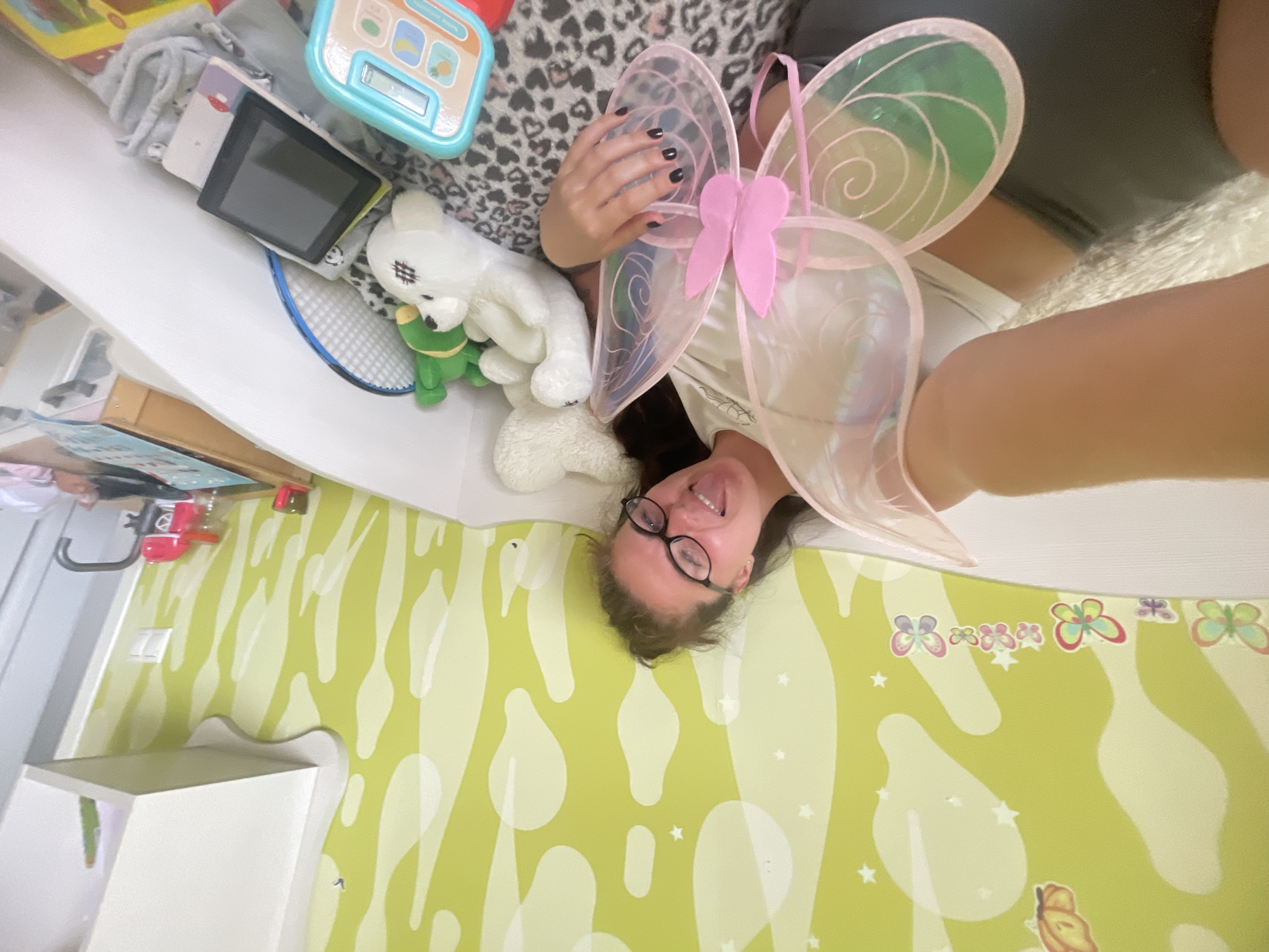 A person with dark hair and glasses is lying on their back on a surface covered in yellow and white patterned wallpaper with butterfly stickers. The person is wearing a pair of pink butterfly wings. They are sticking their tongue out and smiling. A shelf with assorted items is above the person. The items include a tablet, toys, and a white stuffed bear. The location appears to be indoors, possibly a child's room, but the exact city (Moscow, Russia) cannot be determined from the image.