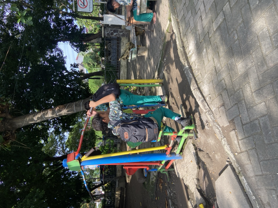 The scene is a public park with a playground in the background. The image shows a young boy using an outdoor exercise machine. He is wearing a black backpack, a multi-colored shirt, and green pants. He is using his arms to pull on a metal bar attached to the exercise machine. The machine is colorful with red, blue, green, and yellow elements. Another young boy in a green shirt, black pants, and a backpack is standing beside the machine, watching the first boy. A third boy is seated on a nearby bench to the right of the image, looking at his phone. The location is in the city of Kecamatan Sidoarjo, Indonesia, at coordinates (latitude: -7.441, longitude: 112.705). The scene is surrounded by trees and there is a concrete path. The ground is covered in dirt and grass. There is a smoking no-smoking sign in the top right corner of the image. The boys seem to be enjoying their time in the park.