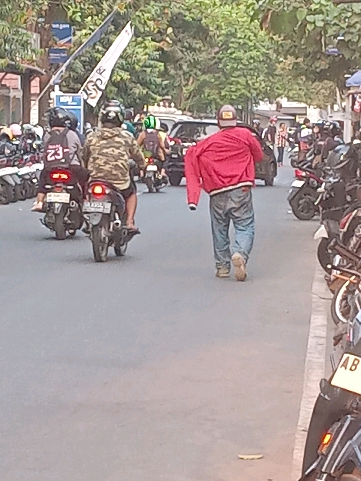 A man in a red jacket and blue jeans walks away from the camera on a street in Yogyakarta, Indonesia. He is carrying his red jacket over his left arm. In the background, several motorcycles are parked on the right side of the street. On the left side of the street, a group of people on motorcycles are moving away from the camera. The street is paved and lined with trees.