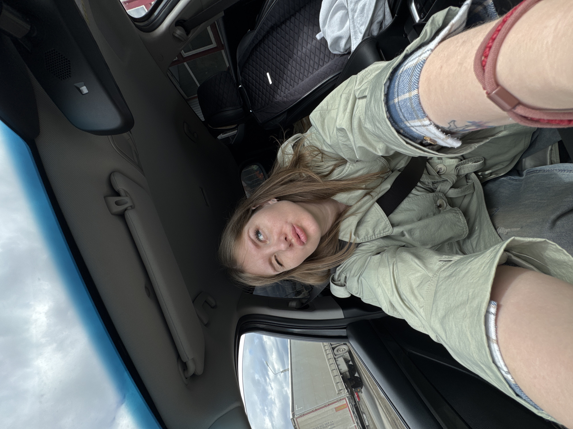 A young woman is captured in a diagonal selfie taken from inside a car during the daytime. She has long, light brown hair and light-colored eyes, looking towards the camera with a playful, slightly pursed-lip expression. She is wearing a light olive green, button-up jacket or shirt over what appears to be blue and white plaid shorts or a skirt. A faint tattoo is visible on her upper left arm, and a reddish-brown hair tie adorns her wrist. A car seatbelt crosses diagonally over her torso.
The interior of the vehicle is visible, including the rearview mirror with a speaker grille and a small button, a sun visor, and part of the car's ceiling. A dark, quilted bag or cushion rests on the seat beside her. Through the car's side window, a large white and grey truck trailer is seen, suggesting the car is either parked or moving slowly alongside it. The side of the truck trailer displays text, including what appears to be a phone number: "7 495 030 69 90". A cloudy sky is visible through the car windows, indicating overcast daylight. The scene takes place in Perm, Russia.