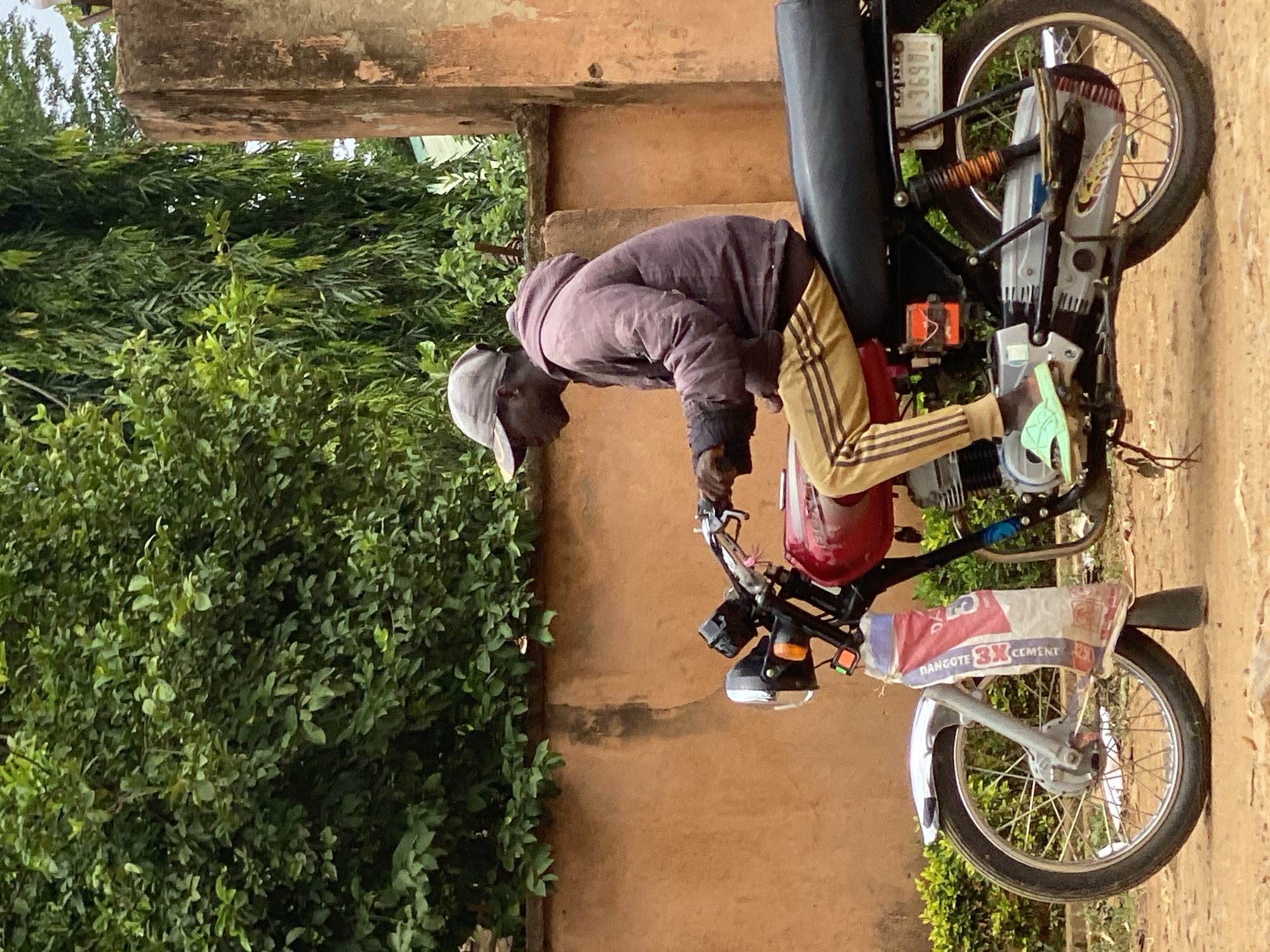 A man wearing a grey cap, dark hooded jacket, and striped pants bends over a motorcycle in Maigizo, Nigeria. The motorcycle has a red fuel tank and a bag labeled "Dangote 3X Cement" attached to the front. The background shows a wall and green foliage. A license plate with "KANO" and "1-3697" is visible on the motorcycle. The scene appears to be outdoors.