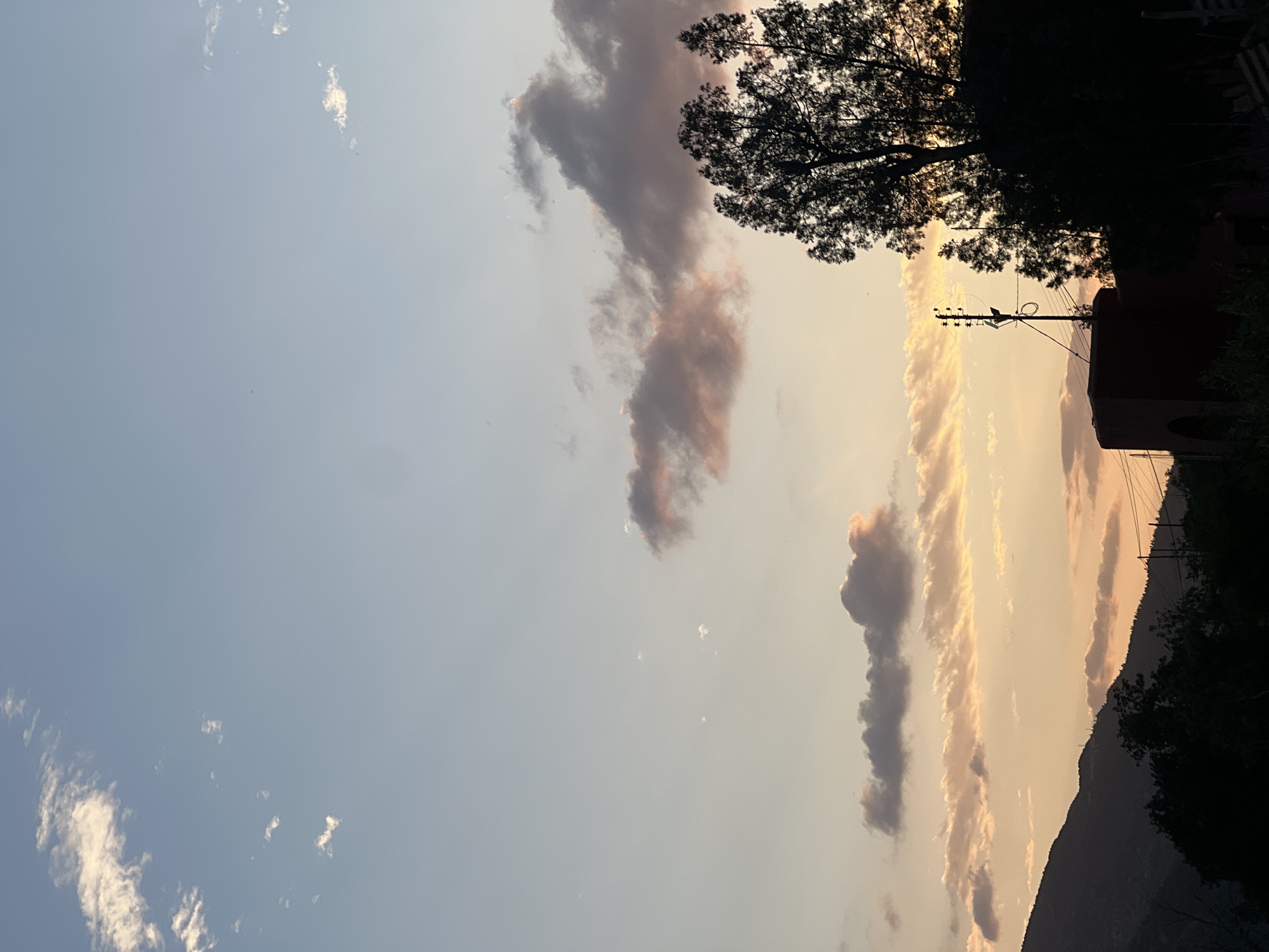 The image shows a landscape view in Bin El Ouidane, Morocco, likely taken during sunset or sunrise. The sky transitions from a pale blue on the left to a warm yellow-orange towards the right horizon. Wispy cirrus clouds are scattered in the blue part of the sky, while more dense, vertical formations of clouds tinged with pink and grey are positioned closer to the right. The silhouettes of trees, a building, and a utility pole with wires are visible against the lighter sky on the right side, indicating the presence of vegetation and infrastructure. The darker silhouette of a mountain range is seen along the bottom-right edge of the image.