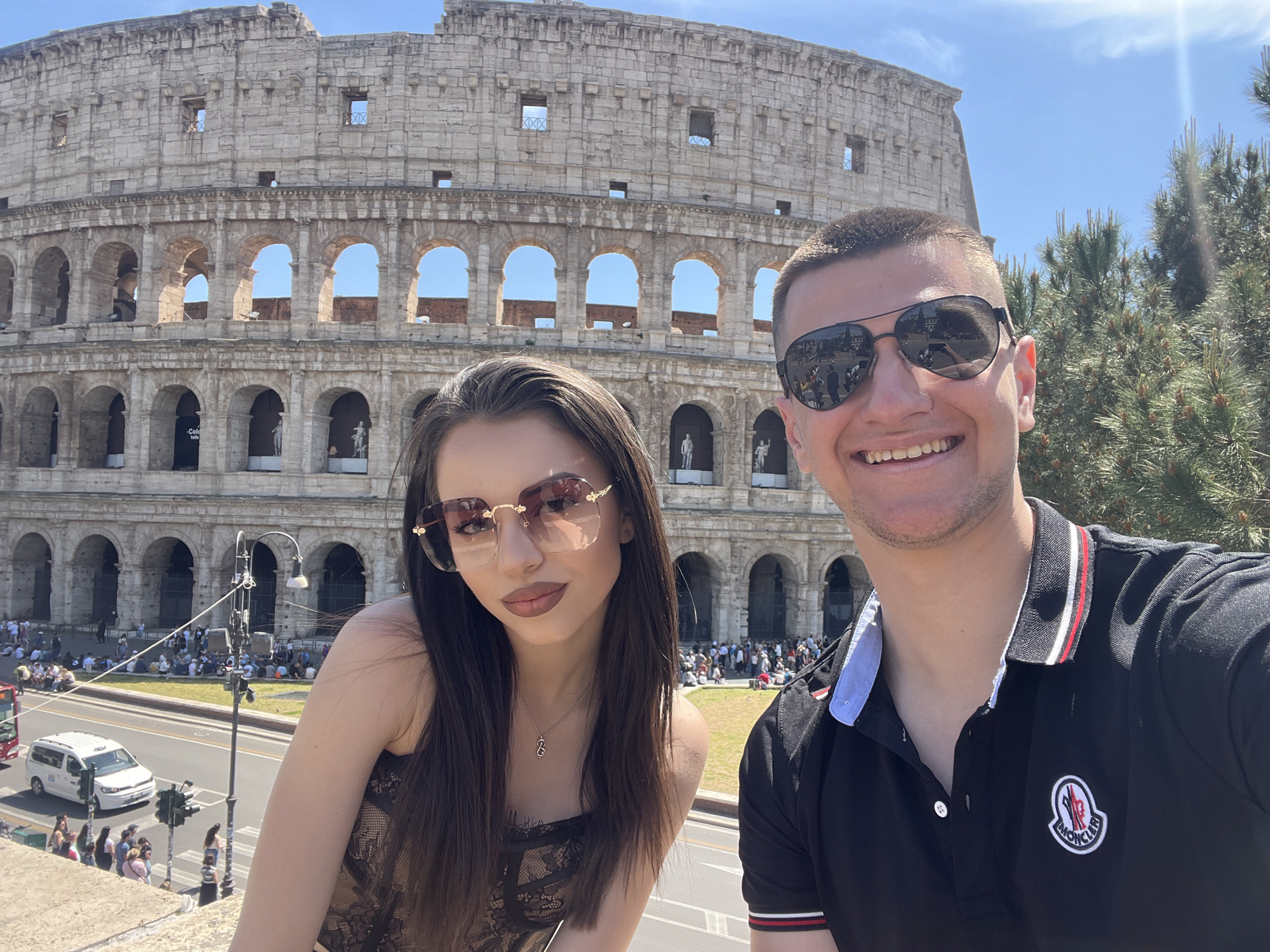 A vibrant outdoor selfie captures a smiling couple posing in front of the iconic Colosseum in Rome, Italy, under a clear blue sky. The bright sunlight suggests it's daytime.
On the left, a woman with long dark hair wears stylish, ornate sunglasses with reddish-brown tinted lenses. She is dressed in a black, lace-patterned top or dress and a gold necklace featuring a distinct 'E'-shaped pendant. She looks towards the camera with a subtle smile.
To her right, a man with short dark hair smiles broadly. He wears aviator-style sunglasses that reflect the surrounding scene, including people and buildings. His black polo shirt has a white collar lining with red and white stripes on the collar and sleeve cuffs, and a prominent white and red Moncler logo is embroidered on the left chest.
The majestic stone structure of the Colosseum serves as the background, displaying its characteristic multiple tiers of arches and weathered facade. Through some of the arches, glimpses of the bright blue sky are visible, and a banner inside one arch partially shows the text "Colo..." (likely "Colosseum tickets"). Below the ancient monument, a busy street is visible with vehicles, including a white van and a red bus, alongside numerous tourists gathered on foot and in grassy areas. A street lamp is also present in the middle ground. The overall scene conveys a cheerful and bustling atmosphere at a famous historical site.