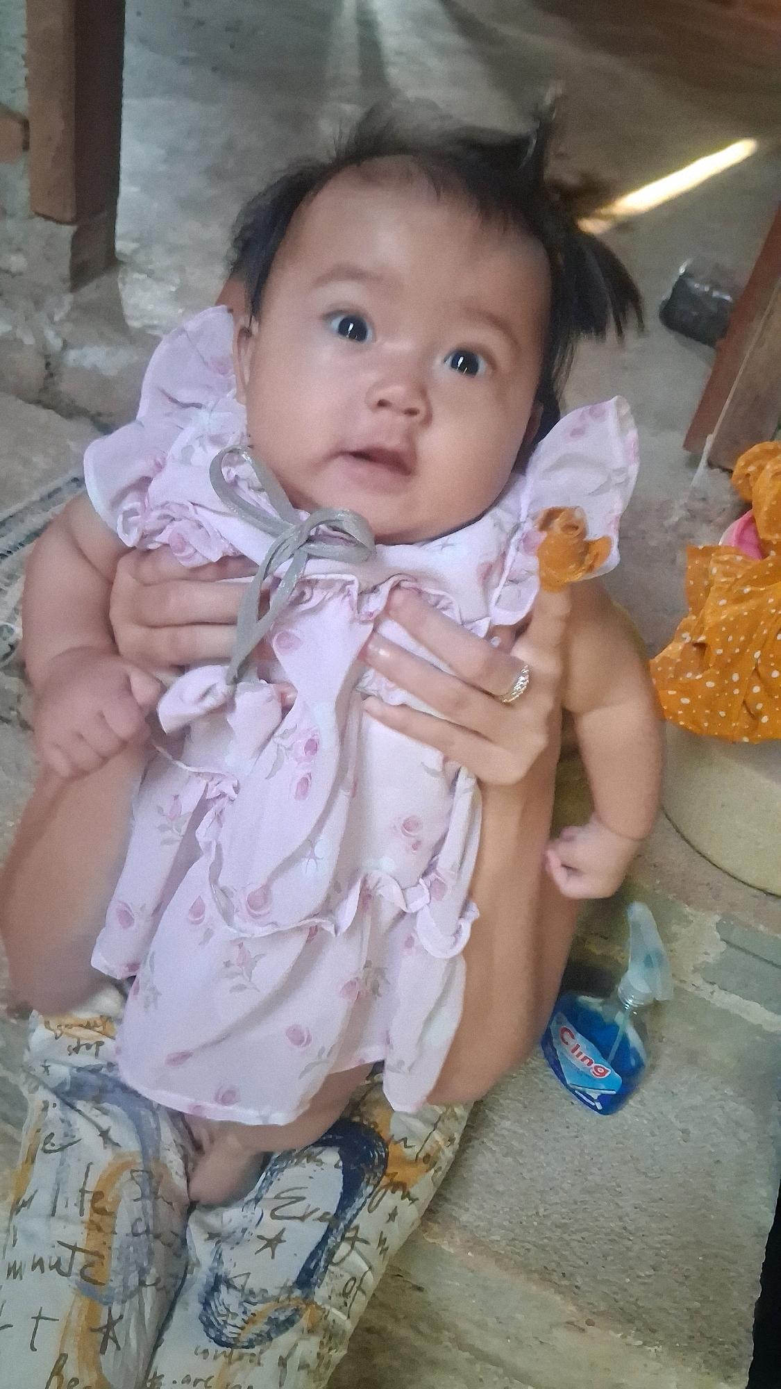 A baby is being held in a seated position, possibly on the floor, as indicated by the rough, textured ground which could be concrete or stone. The baby is wearing a light pink dress with a floral pattern and ruffled sleeves, tied with a gray ribbon. The baby's head is tilted slightly, with wide, dark eyes looking towards the camera and dark, somewhat messy hair. The hands holding the baby are visible, with one hand displaying a ring on its finger. In the background, there are hints of furniture, possibly a table leg and a wooden structure, and on the right side of the image, a brightly colored orange fabric with polka dots is visible, and a blue spray bottle labeled "Cling" is on the ground. The lighting suggests an indoor or covered outdoor setting, possibly in Karang Sari, Indonesia, given the general appearance of the surroundings.