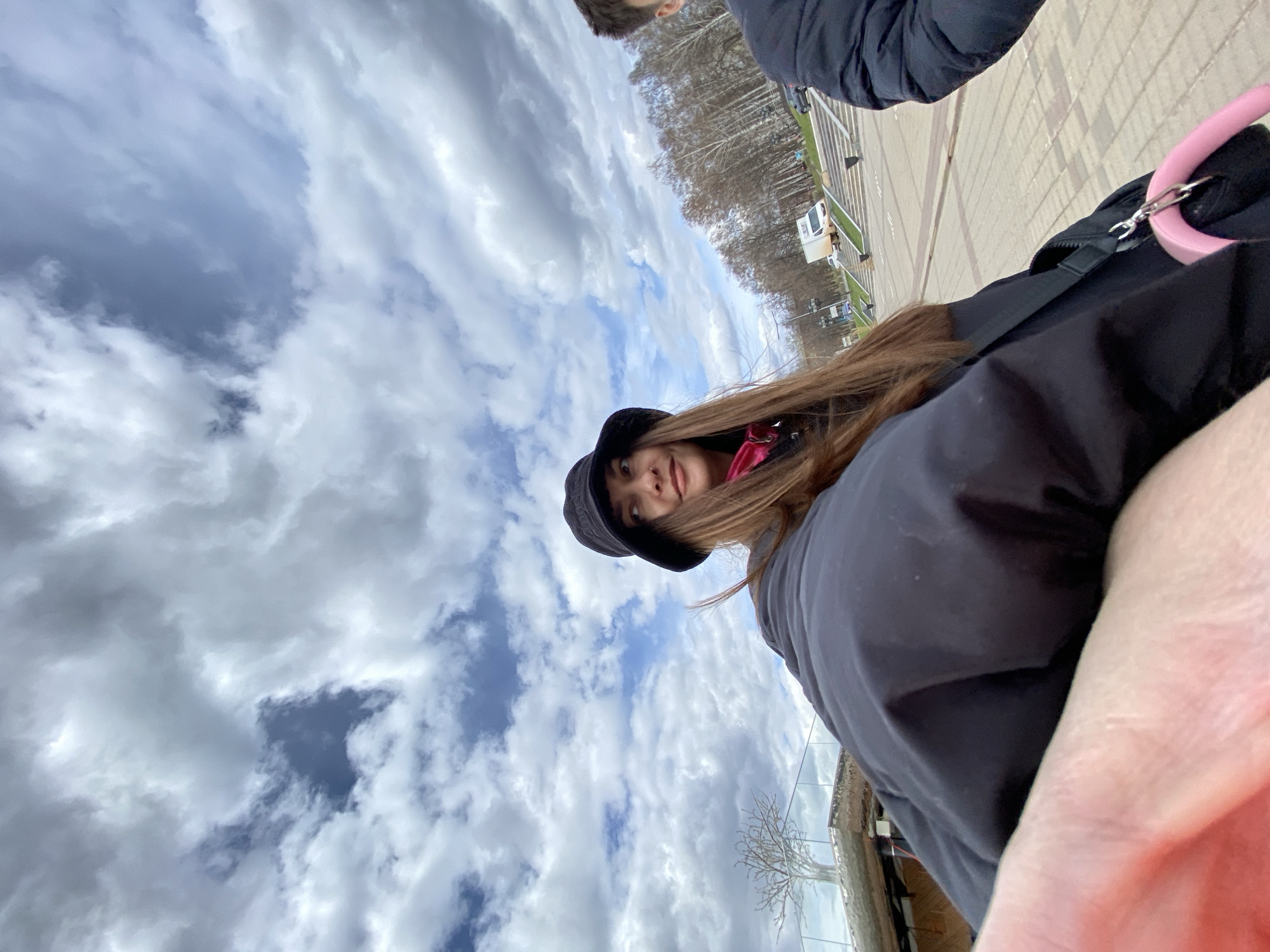 The image shows a selfie of a young woman in Votkinsk, Russia. She's wearing a black bucket hat, a black jacket, and a pink scarf. The sky is cloudy, and there is a road and some trees in the background. Other people are visible in the background. The environment appears to be an urban park or street.