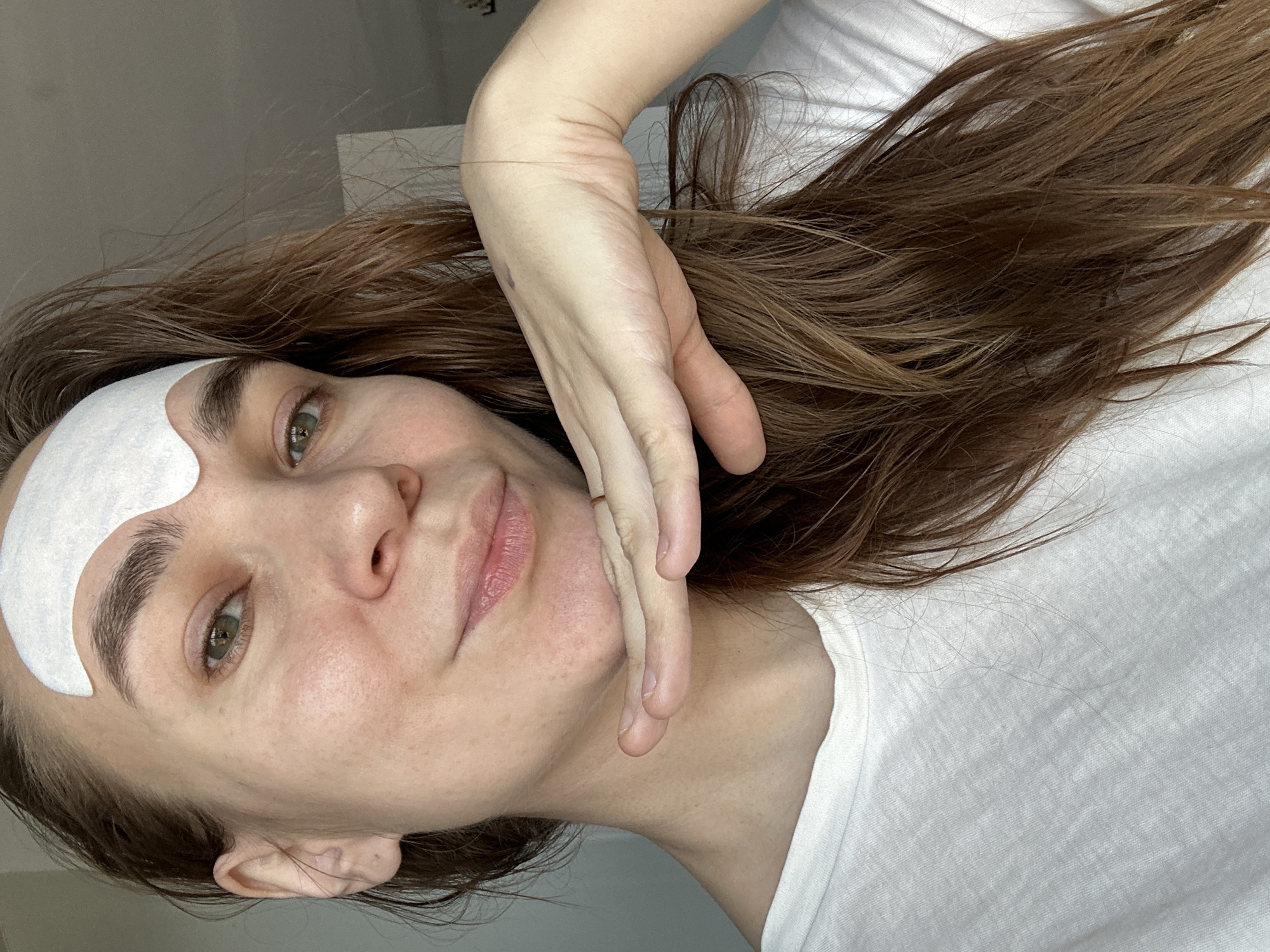 A woman with brown hair is looking at the camera. She is wearing a white t-shirt and has a white patch on her forehead. Her hand is resting on her chin. The background is out of focus. There is no information available about the location being Kharnavy, Russia in the image provided.