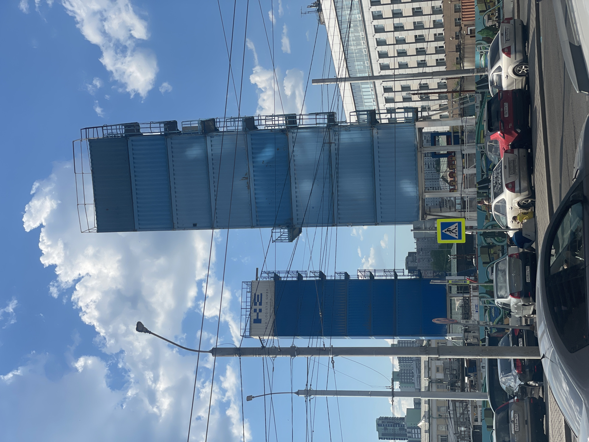 The image depicts a street scene in Krasnoyarsk, Russia. There are several parked cars along the side of the road. Above them are stacked shipping containers, possibly used for a construction or advertising structure. Power lines crisscross the sky. A pedestrian crossing sign is visible. In the background, apartment buildings line the street. The sky is bright blue with scattered clouds. The overall impression is an urban landscape with elements of both residential and industrial character.