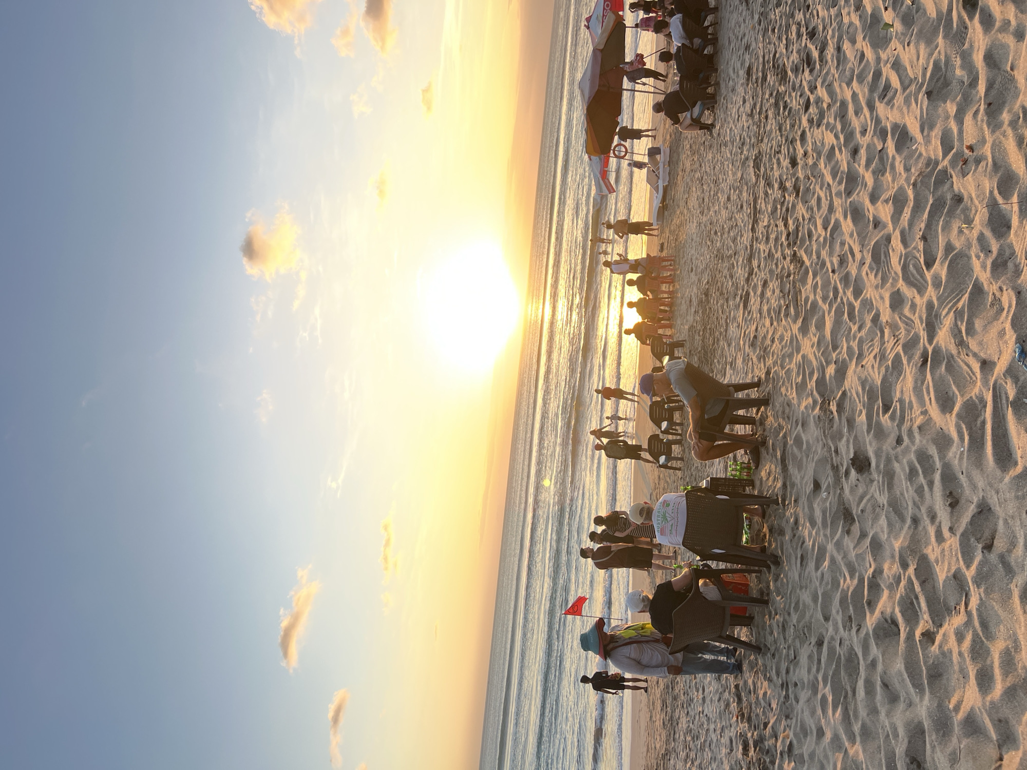 A beach scene at sunset in Kuta, Indonesia, features a group of people relaxing on the sand. The sun, a bright yellow orb, is setting over the ocean, casting a warm glow across the sky and water. Several individuals are seated in chairs, some with drinks, facing the ocean. Others are standing or walking along the shore. The beach is sandy, with small waves lapping at the edge of the water. Beach umbrellas and lounge chairs are visible in the background. A red flag is planted in the sand near a group of people. The overall atmosphere is one of leisure and relaxation.