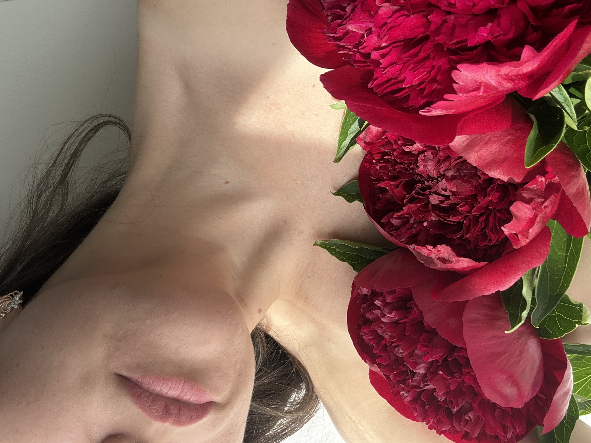 This close-up, artfully composed portrait features a person, likely female, reclining with vibrant red peonies. The subject's face is partially visible, showing her soft pink lips, chin, and cheek, along with an intricate, gold-colored earring with a floral design on her ear. Her long, dark brown hair cascades around her head and neck.
The focus is largely on the elegant curve of her neck and shoulder, which serves as a canvas for three large, lush red peonies with green leaves. The flowers are arranged diagonally across the upper right portion of the frame, contrasting beautifully with the fair skin of the subject. Subtle freckles or moles are visible on her neck and chest area, adding natural detail to her skin.
The lighting is soft and even, suggesting an indoor setting, possibly using natural light or studio illumination, creating a serene and delicate mood. There is no specific activity depicted beyond a posed portrait. No text is visible in the image. This photographic artwork was captured in Chaikovsky, Russia.