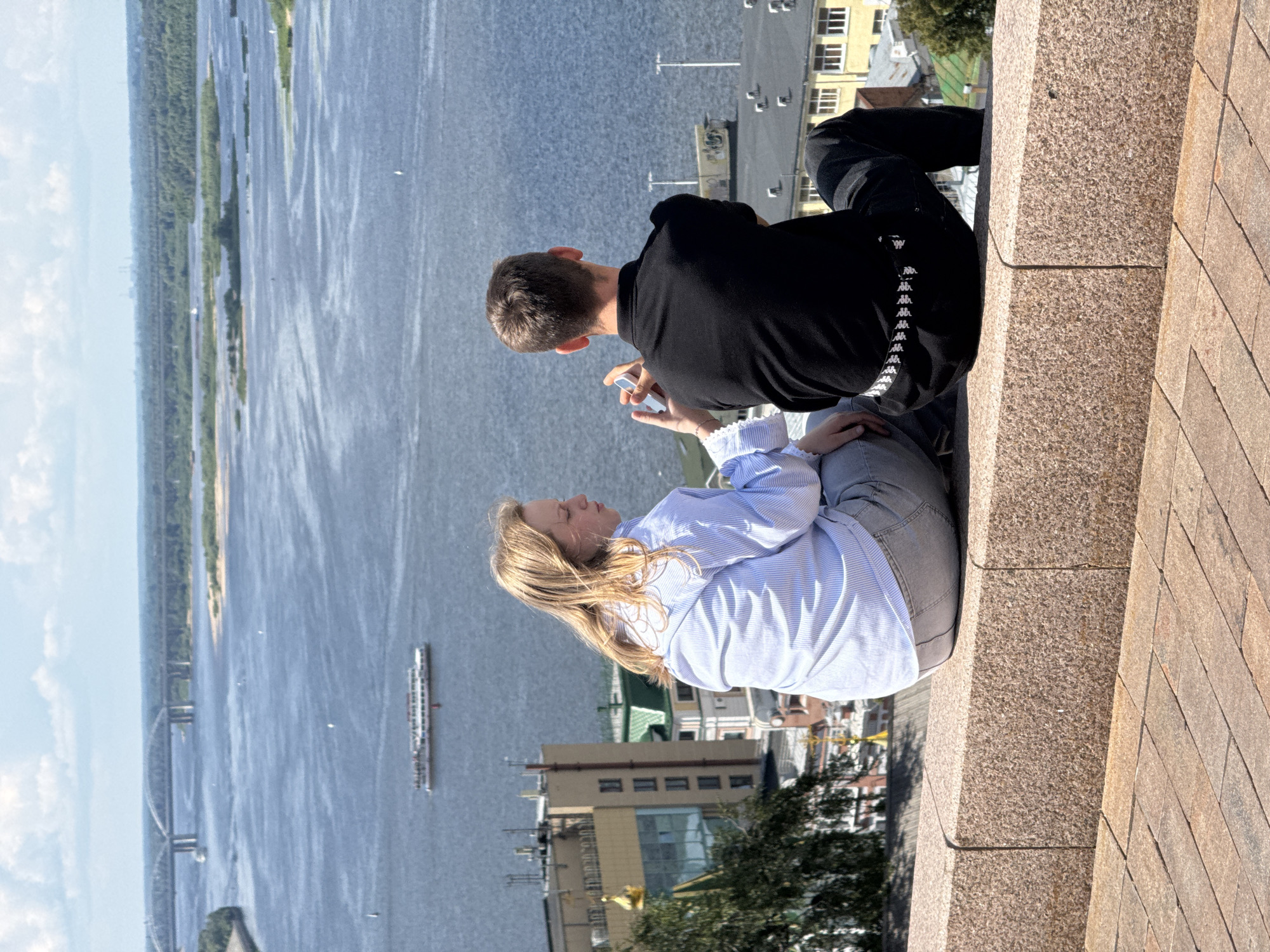 Two young people, a girl with blonde hair and a boy with brown hair, are sitting on a granite ledge overlooking a wide river and a cityscape. The girl is wearing a light blue and white striped shirt and grey pants, and she is holding a phone in her hands, as if taking a picture. The boy is wearing a black polo shirt and black pants, and he is looking out at the river. The river is wide and blue, with a few boats on it. In the background, there are bridges, buildings, and trees. The sky is partly cloudy. The scene appears to be from a viewpoint in Nizhny Novgorod, Russia, likely from a high point such as the Kremlin or one of the city's many scenic overlooks, given the panoramic view of the Volga River and the urban landscape.