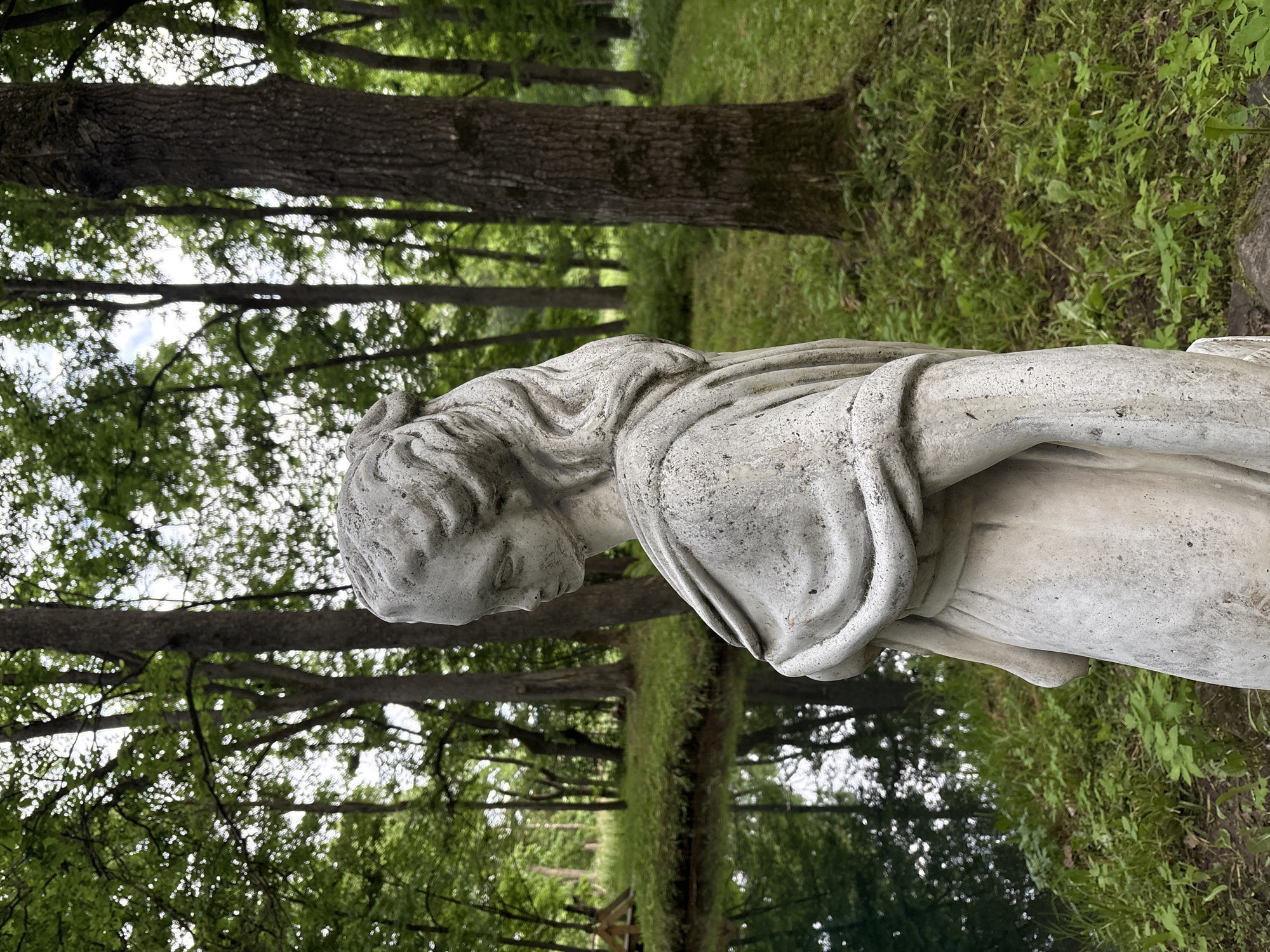 The image is an outdoor shot of a stone statue of a woman in a garden setting. The statue is in the foreground, facing left in profile, and is weathered with visible texture. It depicts a woman with her hair styled up and wearing a draped garment. Behind the statue, there is a body of dark water, possibly a pond or small lake, with reflections of trees. The background consists of a dense forest with tall trees and green undergrowth. The overall scene suggests a natural, perhaps slightly overgrown, park or garden environment. There are no people or specific landmarks that would indicate the city of Gorki, Russia.