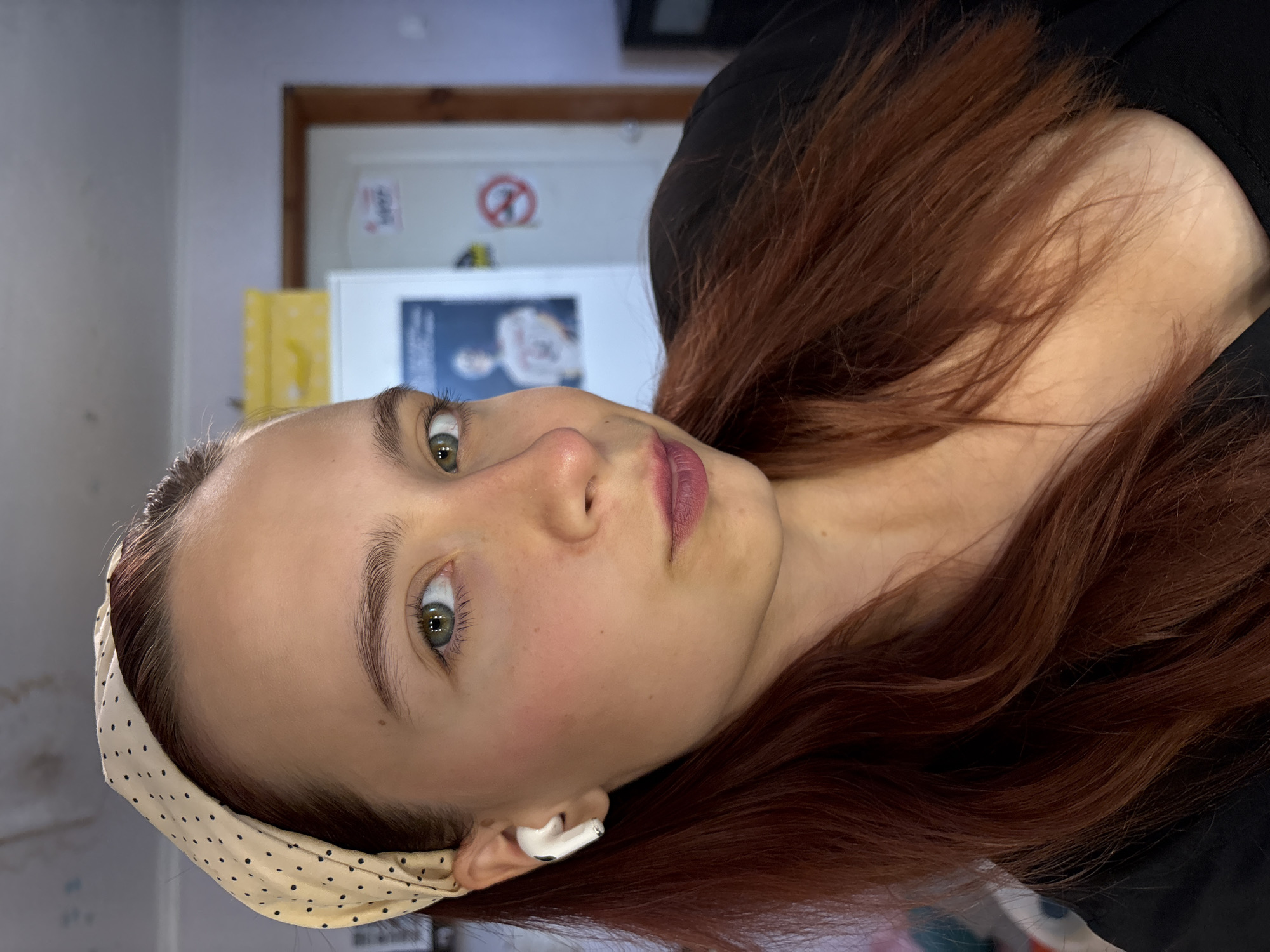 A young woman with auburn hair and green eyes is pictured in a close-up, head-and-shoulders shot. She is wearing a beige headband with black polka dots and a white wireless earbud in her left ear. Her makeup includes blush on her cheeks and dark pink lipstick. She is looking directly at the camera with a neutral expression. Her hair is long and wavy, falling over her shoulders and chest. She is wearing a black garment. In the background, a white refrigerator, a doorway with a wooden frame, and a wall painted in a light purple hue are visible. There is a picture on the refrigerator door and a red and white "no smoking" sign on the wall to the right of the doorway. The overall environment suggests an indoor residential setting. The image does not provide any specific visual cues to pinpoint its location within the city of Прикамский, Russia.