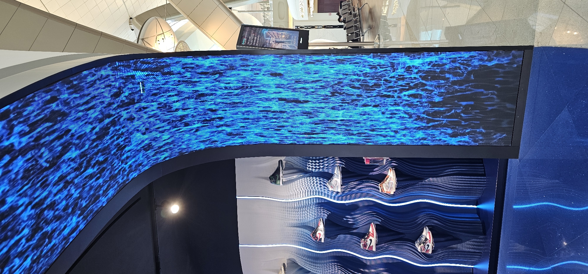 The image shows an indoor display area with a curved LED screen displaying a blue, abstract pattern resembling flowing water or flames. To the left of the LED screen is a display of shoes on elevated shelves against a wavy, textured backdrop with vertical blue lighting. The floor is a reflective dark blue. In the background, beyond the display, an airport terminal environment is visible with seating areas, a digital advertisement screen, and architectural elements characteristic of an airport, such as large ceilings and floor tiling. The location is identified as Al Matar, United Arab Emirates. There are no discernible people in the foreground, but figures are visible in the background seating area.
