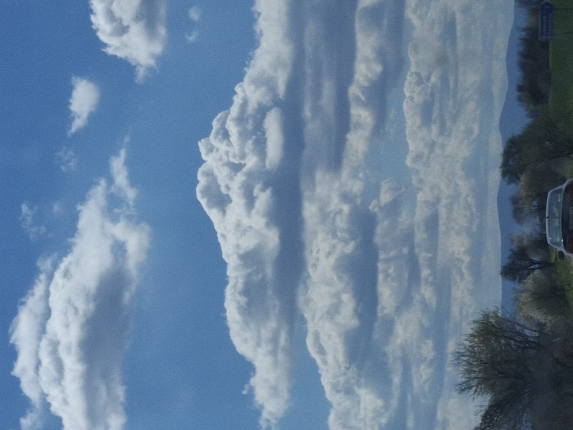 The image shows a view of the sky with various cloud formations. There are fluffy, cumulus-like clouds in the blue sky. Near the horizon, there are trees and glimpses of a building and vehicle. This is probably in Gramada, Bulgaria.
