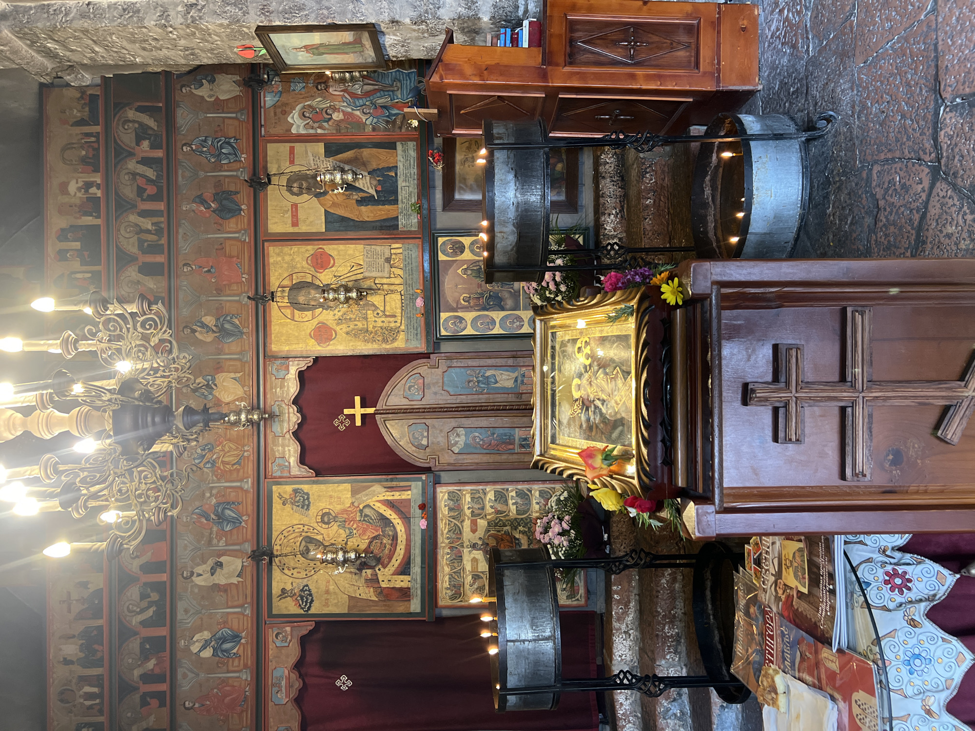 This image shows the interior of the Church of St. Elijah, located in Petrovac centar, Montenegro. The walls are covered in religious iconography and artwork, with several icons displayed prominently. A chandelier hangs from the ceiling, illuminating the scene. Candles are lit in metal holders. A wooden lectern or desk is also visible. A cross is displayed on a wooden panel. Various religious items and books are arranged in the space. The environment suggests a place of worship and religious significance.