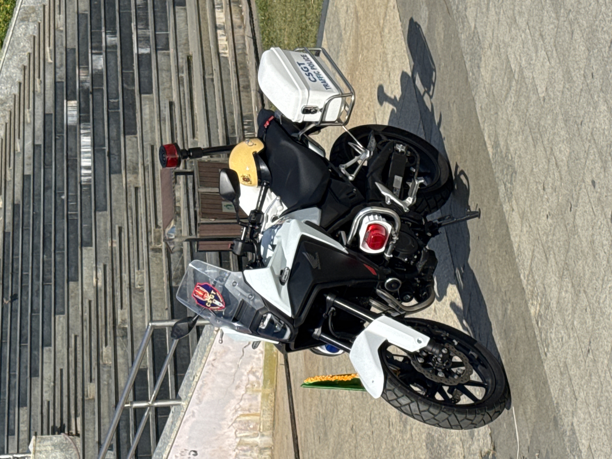 A stationary, white Honda motorcycle, equipped for traffic police duty, is parked on a paved walkway in front of a wide, multi-tiered stone staircase. The motorcycle features a prominent windshield with a circular red, white, and blue emblem. A yellow helmet rests on the seat, and a red flashing light is mounted on a black pole behind the handlebars. A white storage box, labeled "CSGT TRAFFIC POLICE," is attached to the rear of the motorcycle. A green and yellow garland is visible to the left of the front wheel. The background consists of the textured stone stairs and a wall with peeling paint. The lighting suggests it is daytime, with shadows cast on the ground. The scene does not depict any people or ongoing actions.