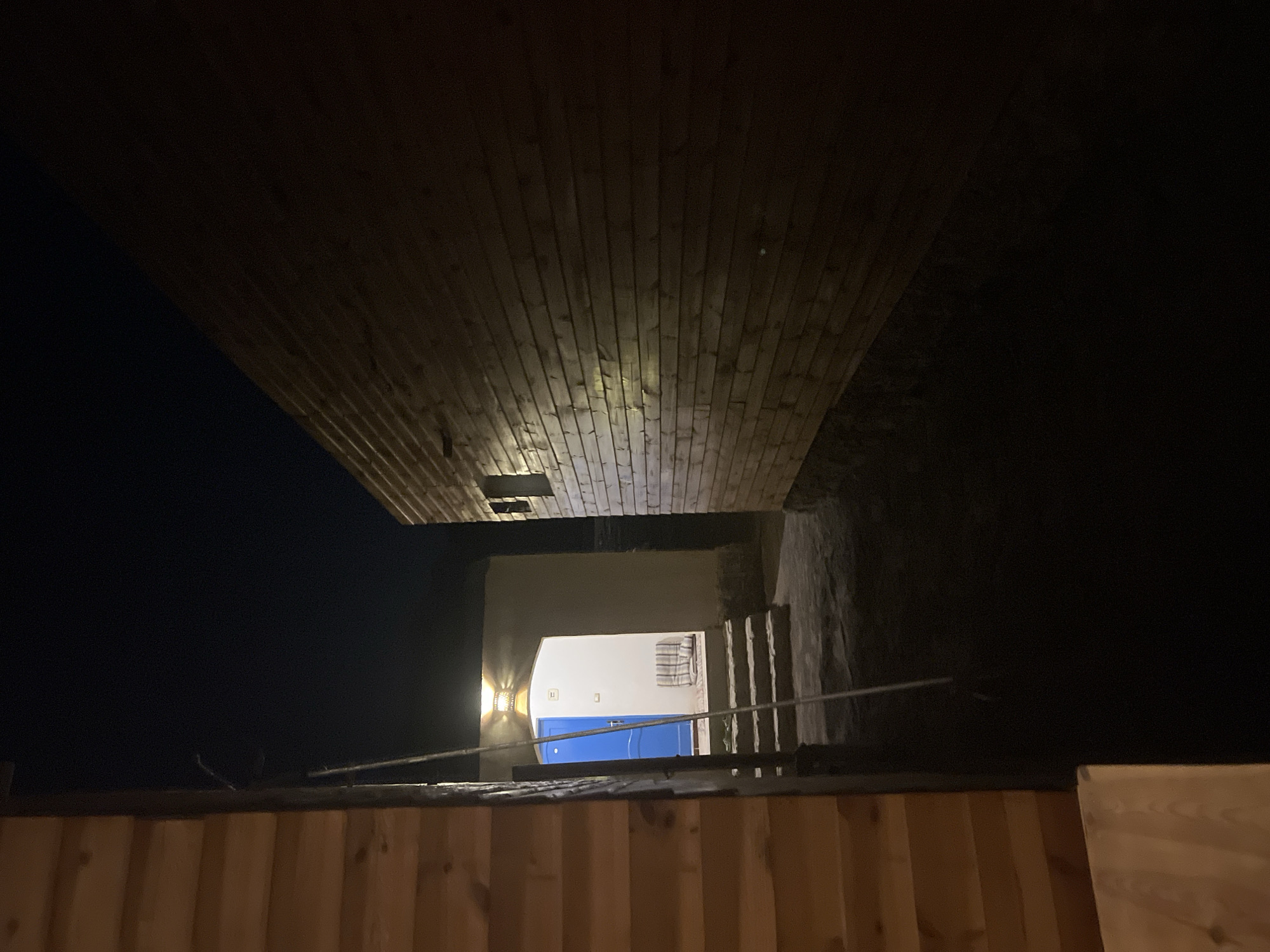 A pathway at night leads to a brightly lit doorway with a blue door, situated between two wooden structures in the city of Nueiba, Egypt. The pathway is lined with stone steps and a rope hangs down from above on the left side of the image. One wooden structure is on the left, partially obscuring the view, and appears to be a building with horizontal wooden planks. The other wooden structure is on the right, with the same planking style and seems to be a wall. The doorway ahead is framed by an arch and has a warmly lit lamp above it. Inside the doorway, a white wall is visible, along with a small chair or cushion with a checkered pattern. The sky above is dark, indicating nighttime. The scene suggests an outdoor area, possibly a courtyard or alleyway, within a residential or hospitality setting.
