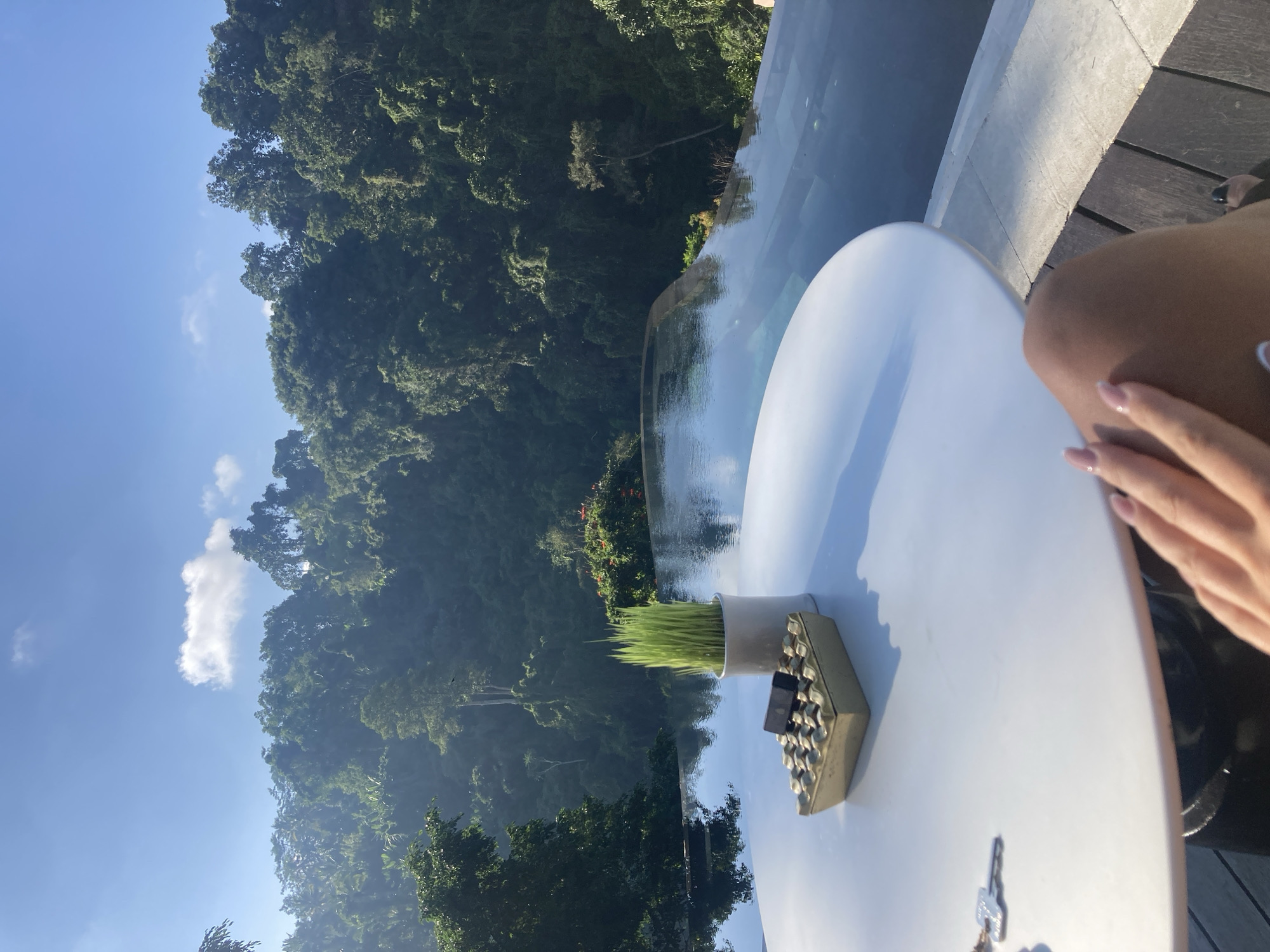 In Payangan, Indonesia, a person's legs and hand are visible in the foreground, resting on a white circular table next to an infinity pool. The table holds an ashtray with a small dark object and a pot with green grass. Beyond the pool, a lush, dense green forest covers rolling hills under a clear blue sky with scattered clouds. The pool's surface reflects the sky and the surrounding greenery, creating a serene and expansive atmosphere.