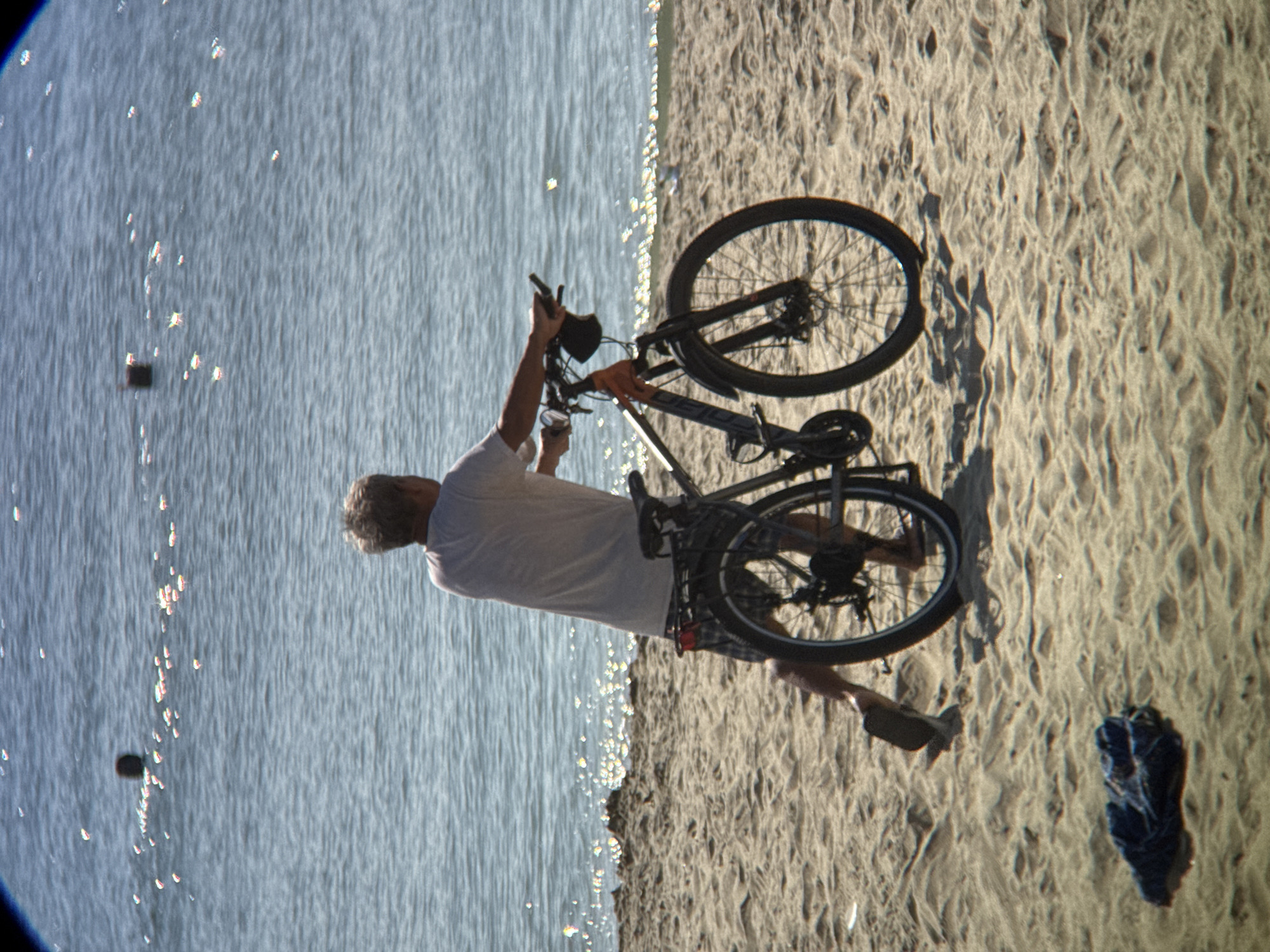 A person, seen from behind, stands on a sandy beach next to a bicycle. The person is wearing a light-colored t-shirt and checkered shorts. Their right leg is extended backwards, and they are holding the handlebars of the bicycle with both hands. The bicycle is dark-colored with orange accents on the frame. The background consists of the calm, blue water of the ocean, with sunlight glinting off its surface. There are a few small, dark objects floating in the water. In the foreground, on the sand to the left of the bicycle, is a crumpled piece of dark blue fabric, possibly clothing. The scene is likely taking place on a beach in Nha Trang, Vietnam, given the calm ocean and sandy terrain.