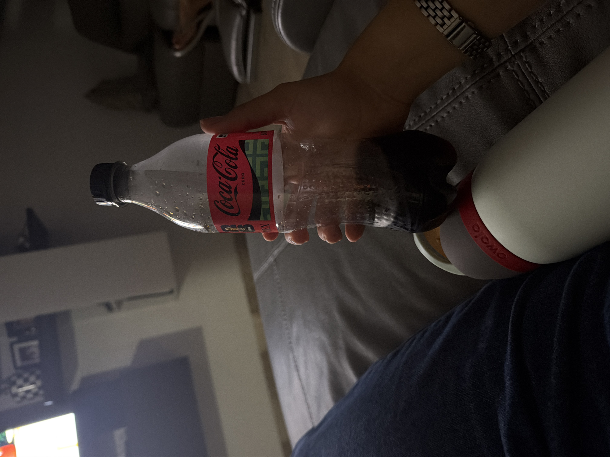 A person is holding a nearly empty plastic Coca-Cola Zero bottle, pouring the remaining liquid into a white Owala water bottle. The scene takes place indoors, likely in a home setting, suggested by the presence of a gray couch, blue jeans, a watch on the person's wrist, and a glimpse of furniture and a television in the background. There is no visible information that indicates the location being Santa Clara Ocoyucan, Mexico.