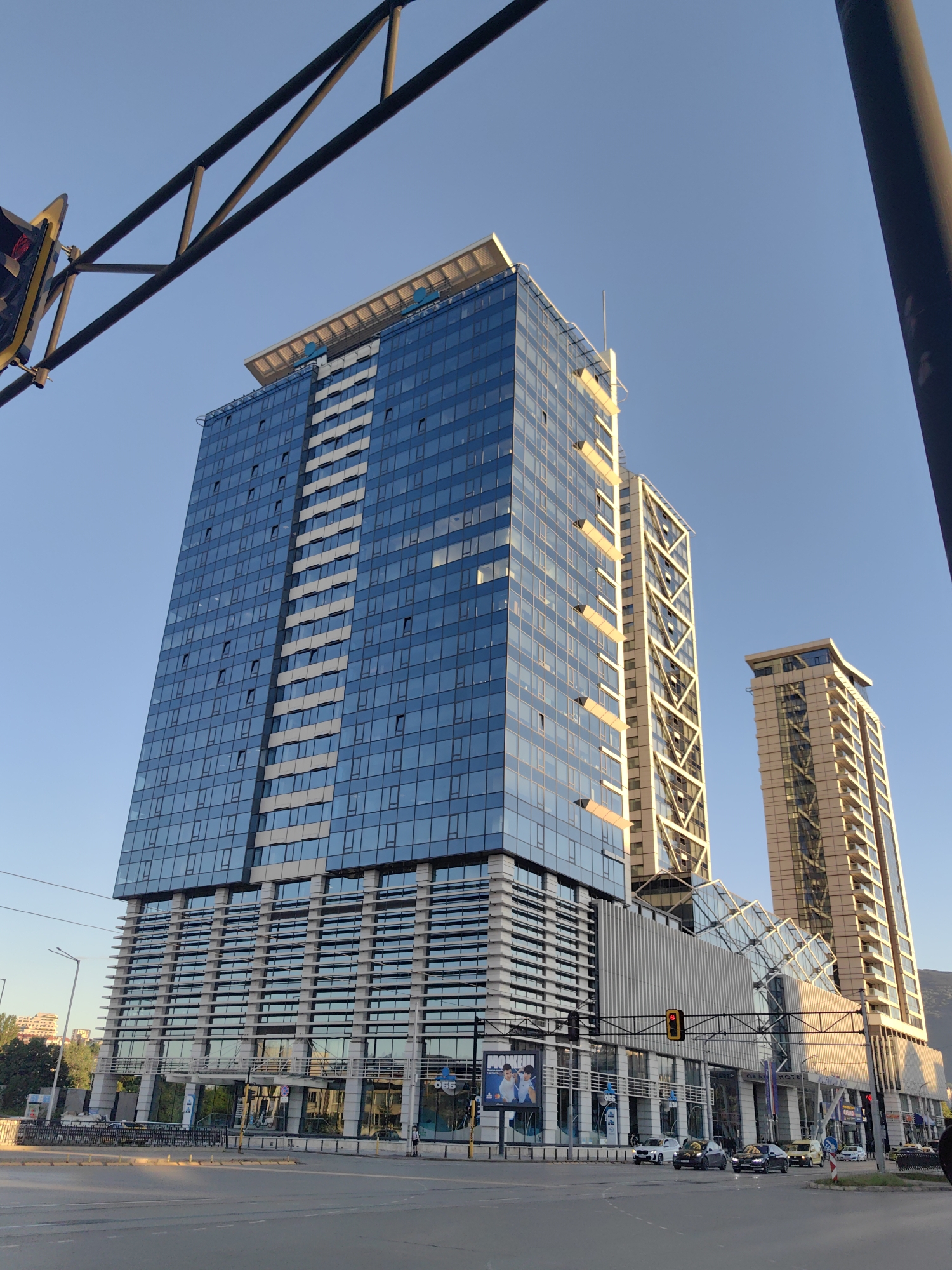 The image shows a view of the Capital Fort office complex in Sofia, Bulgaria. The view is captured from street level, looking upwards towards the buildings. The primary building, a modern glass-fronted skyscraper, dominates the left side of the frame. A second, similarly tall building with a distinctive exterior structural pattern is visible behind and to the right of the first. A third tall building is further to the right and slightly behind the second building. A glass-roofed structure connects the bases of the buildings. A busy street scene is at the bottom of the image. Cars are visible on the road. Parts of traffic light infrastructure and a light pole are on the right and left sides of the image. A billboard is visible on the lower level of the main building. The sky is clear and blue.