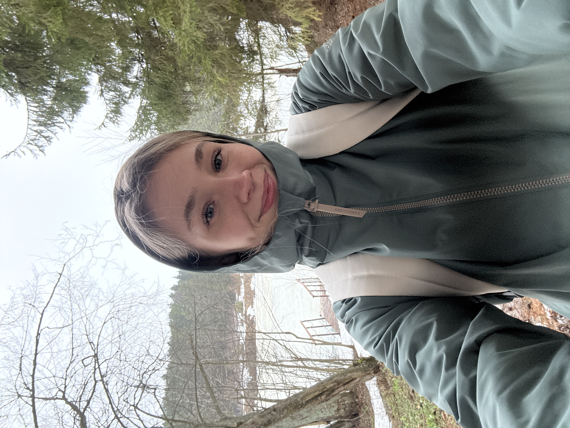 A young woman is taking a selfie outdoors. She is wearing a green hooded jacket and a backpack with beige straps. The background shows a body of water, possibly a lake or river, with bare trees and some greenery. The location appears to be in a forested area. The jacket has the brand name "CORDILLERO" visible on the zipper pull. It cannot be confirmed that the location is in the city of Chaikovsky, Russia based solely on the image.
