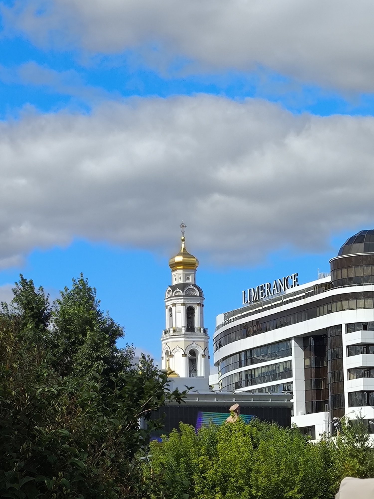 The image captures a scene in Yekaterinburg, Russia, featuring a blend of historical and modern architecture under a partly cloudy sky.
In the foreground, dense green foliage from trees and bushes partially obscures the view. Behind the greenery, a white bell tower with a golden dome, topped with a cross, rises prominently. The tower has arched openings and decorative elements.
To the right of the bell tower, a large, modern, multi-story building is visible. This structure has a curved facade, predominantly white with horizontal bands and dark, reflective windows. A prominent illuminated sign on this building reads "LIMERANCE". A dark, domed section is also visible on the right side of this building.
Below the modern building and partially obscured by trees, a large screen displays a colorful, abstract pattern and what appears to be a statue of a person wearing a hat. The overall impression is a city landscape where older religious structures coexist with contemporary commercial development.