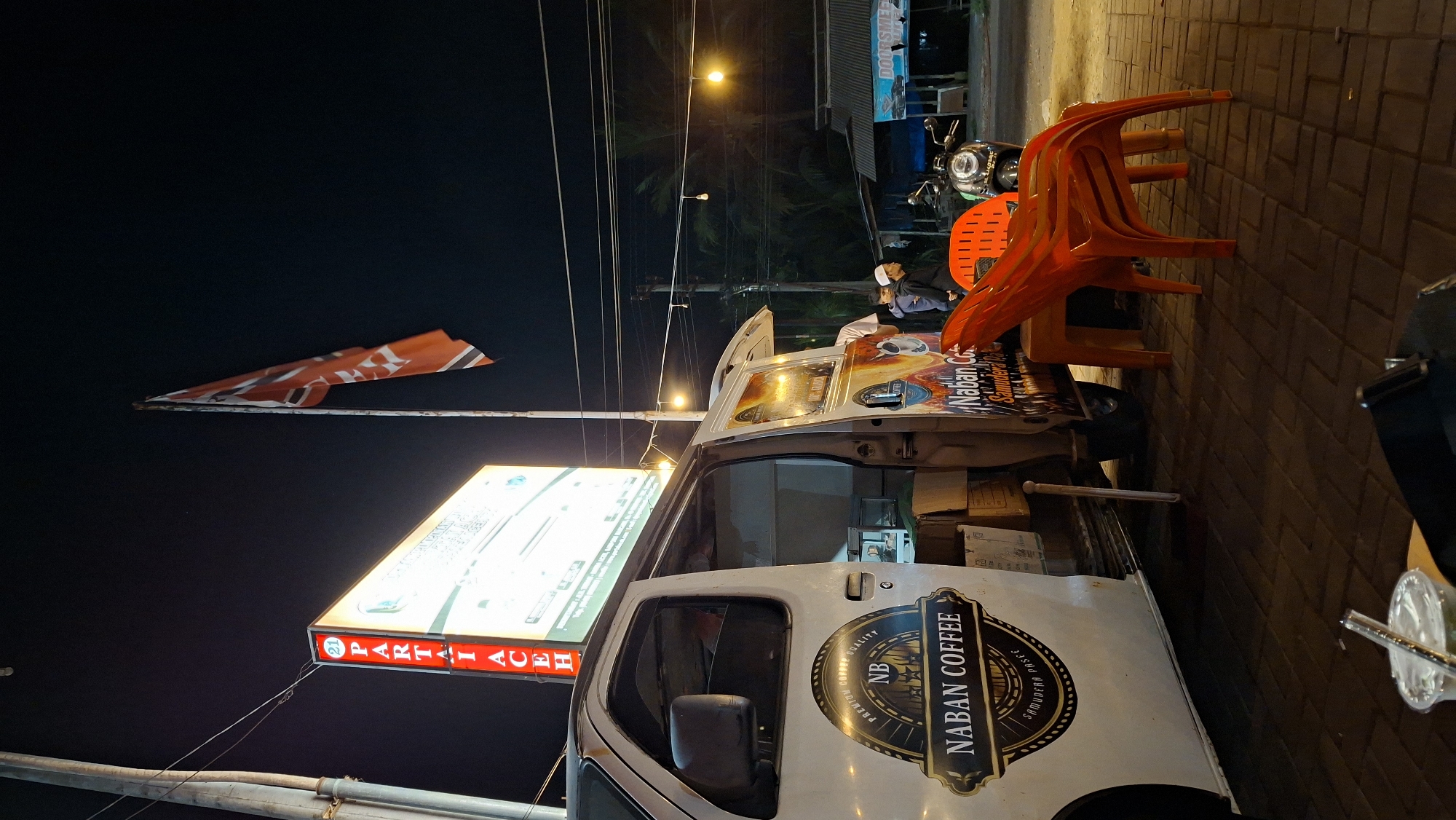 This media file captures a scene at night in Keudeteupinpunti, Indonesia. The primary subjects are a white food truck with "NABAN COFFEE" branding and a large illuminated sign that reads "PARTAI ACEH." A few people are visible, one man wearing a white cap and black jacket, another in a white shirt, and others in the background. The scene is set on a brick-paved street next to the food truck, with an orange plastic chair stacked on top of another in the foreground. Power lines crisscross the dark sky, and streetlights cast a warm glow. A red and white flag is displayed on a pole. The atmosphere appears to be that of a casual evening gathering or a business operation. The visible text "NABAN COFFEE" and "PARTAI ACEH" suggests a coffee vendor operating near a political party's location or event. The time of day is clearly night, and the setting seems to be an outdoor urban or semi-urban area.