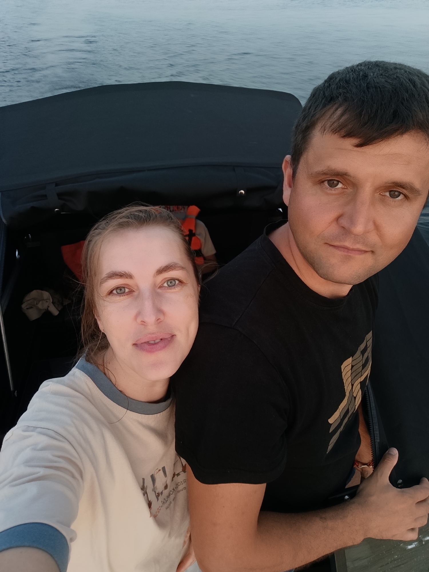 A selfie is taken by a couple on a boat, with the water visible in the background. The woman is on the left, leaning her head on the man's shoulder. She is wearing a light-colored long-sleeved shirt with a blue collar and cuff. The man is on the right, wearing a black t-shirt with a graphic design on it. He is holding onto something dark and textured, possibly the side of the boat. In the background, inside the boat, a life vest is visible. The lighting suggests it is either early morning or late evening. This scene appears to be on the Kama River in the city of Chaikovsky, Russia, given the context of the location.