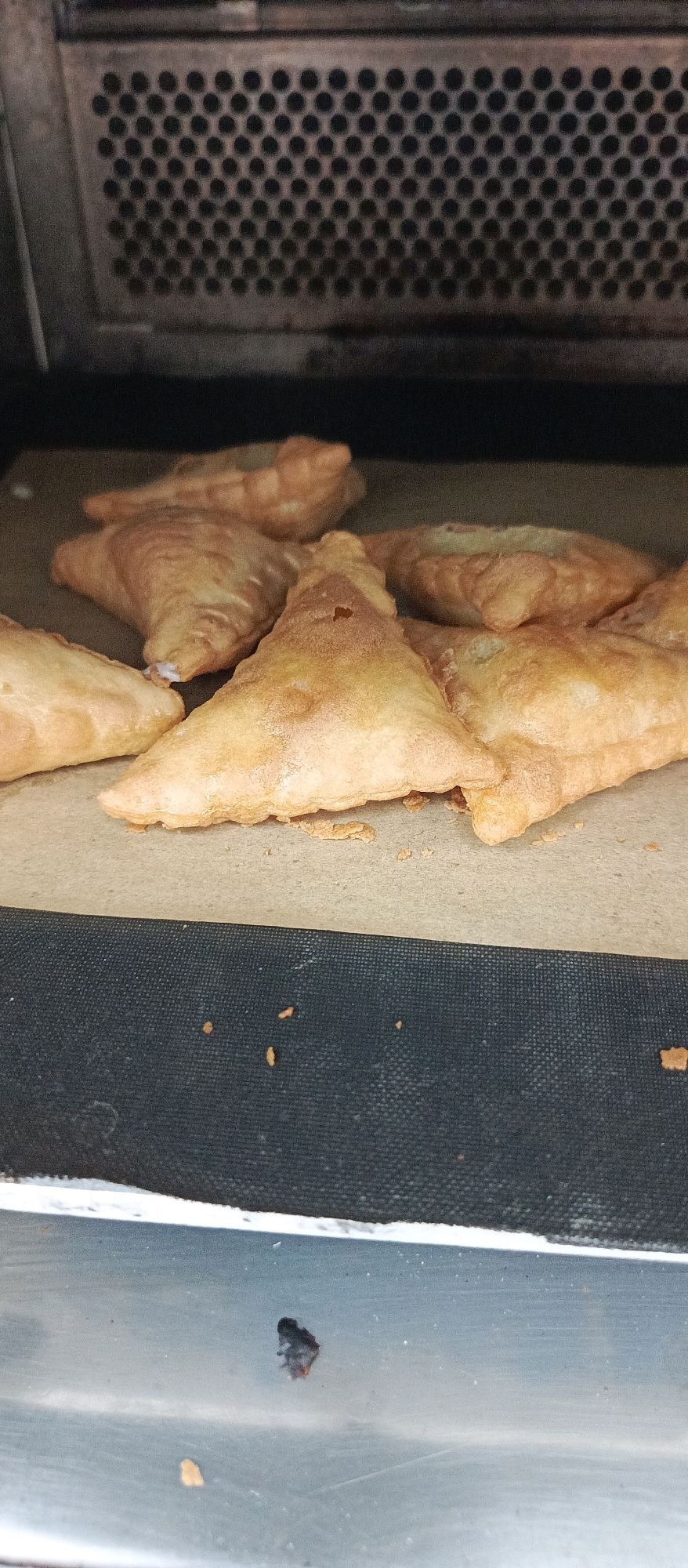 A close-up, low-angle, eye-level shot shows a baking rack inside an oven with several golden-brown, triangular pastries. The pastries appear to be made of a flaky dough and are arranged in a cluster on what looks like parchment paper. The oven interior is visible in the background, with a dark, perforated metal plate. The foreground is occupied by the metallic surface of the oven door or shelf. The lighting is dim, creating soft shadows. The overall scene suggests a homemade baking process. This image was taken in Bugry, Russia.