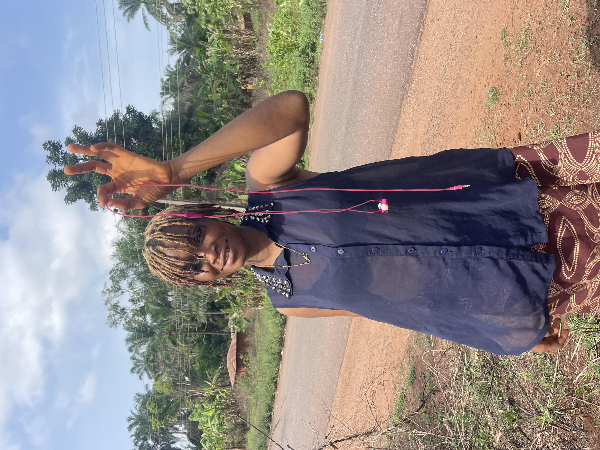 A young woman stands on a dirt and grass shoulder alongside a paved road, seemingly in the outskirts of a tropical area in Ogbe, Nigeria. She is holding pink headphones in her raised left hand. She is wearing a dark, see-through button-down shirt with studs on the shoulders, a gold necklace with an infinity pendant, and patterned brown pants. The background shows a road, lush vegetation, and a cloudy blue sky. Overhead power lines are visible. A small building is visible in the background.