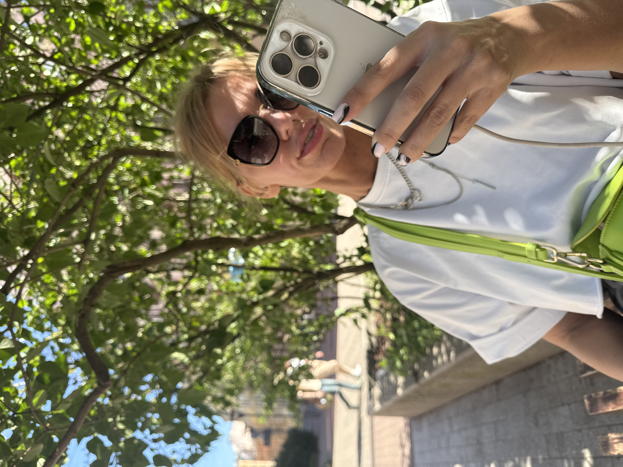 A woman with blonde hair and sunglasses is taking a selfie outdoors. She is wearing a white t-shirt and a lime green purse. Her fingernails are painted black and white. She is holding a smartphone with a prominent camera array. The background shows trees with green leaves, and in the distance, buildings that suggest an urban environment, likely in Saint Petersburg, Russia, given the architectural style and pedestrian activity. The ground appears to be paved with cobblestones. The lighting suggests a sunny day.