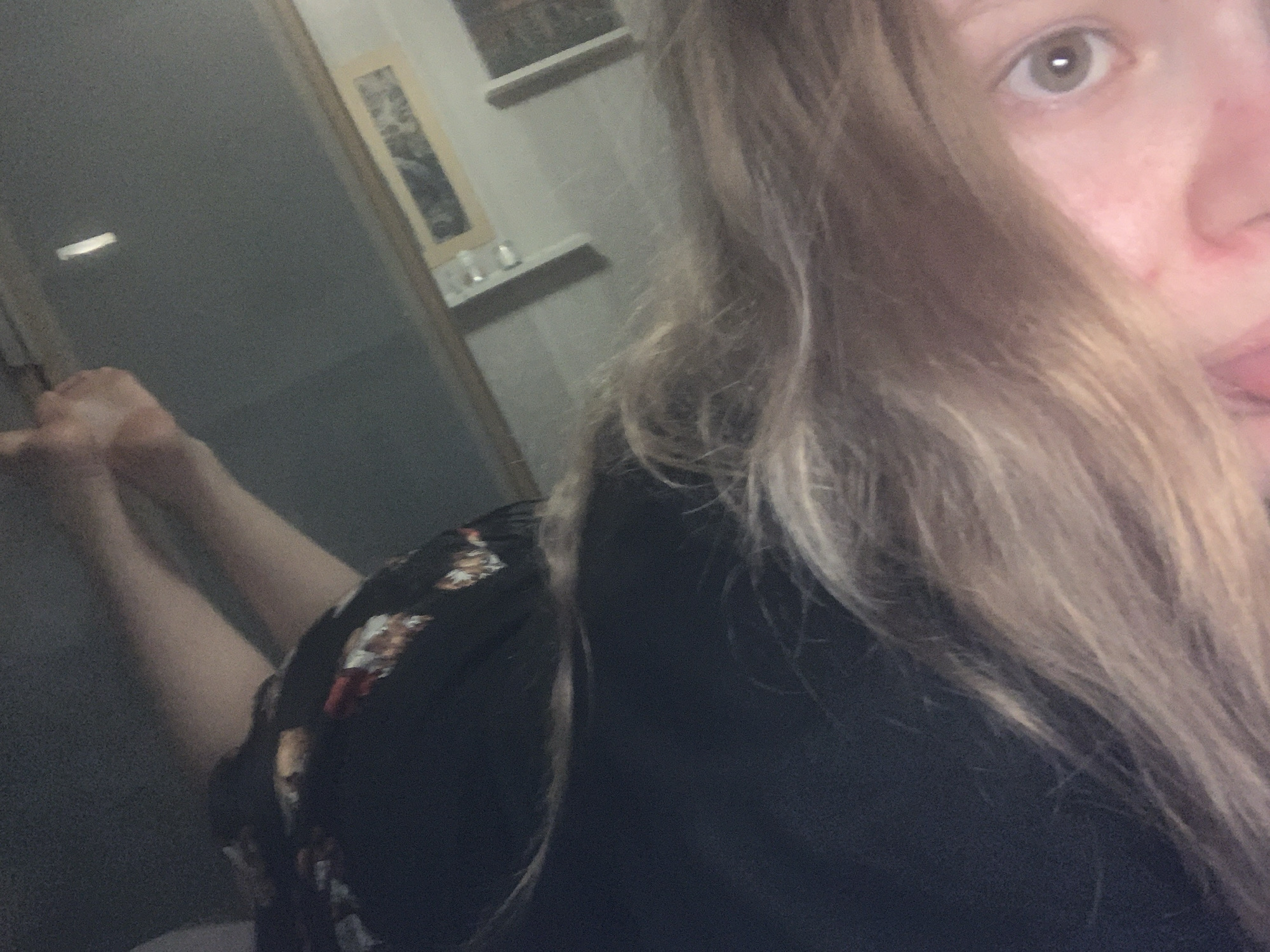 This media file captures a partial view of a person, likely a young woman, in an indoor setting in Vsevolozhsk, Russia. The image is taken from a low, upward angle, possibly a selfie.
The subject's face is prominent in the upper right quadrant, with light blonde or brown hair cascading over much of it. One eye, appearing light in color, is clearly visible, along with the bridge of her nose and part of her mouth where her tongue is slightly extended, suggesting a playful or casual expression. She is wearing a dark, possibly black, top or dress, with a patterned or floral design visible on the left shoulder/sleeve area.
In the mid-left of the image, the person's bare legs are visible, bent at the knees, indicating she might be lying down or lounging.
The background shows an interior room. A light-colored wall is visible, with a framed picture hanging on it. Below the picture, there's a narrow shelf holding a few small, indistinct objects, possibly glasses or bottles. To the far left, a door or door frame is discernible.
The lighting in the scene is somewhat dim, implying it might be evening or an indoor space with subdued illumination. The overall mood appears relaxed and informal. There is no visible text in the image.