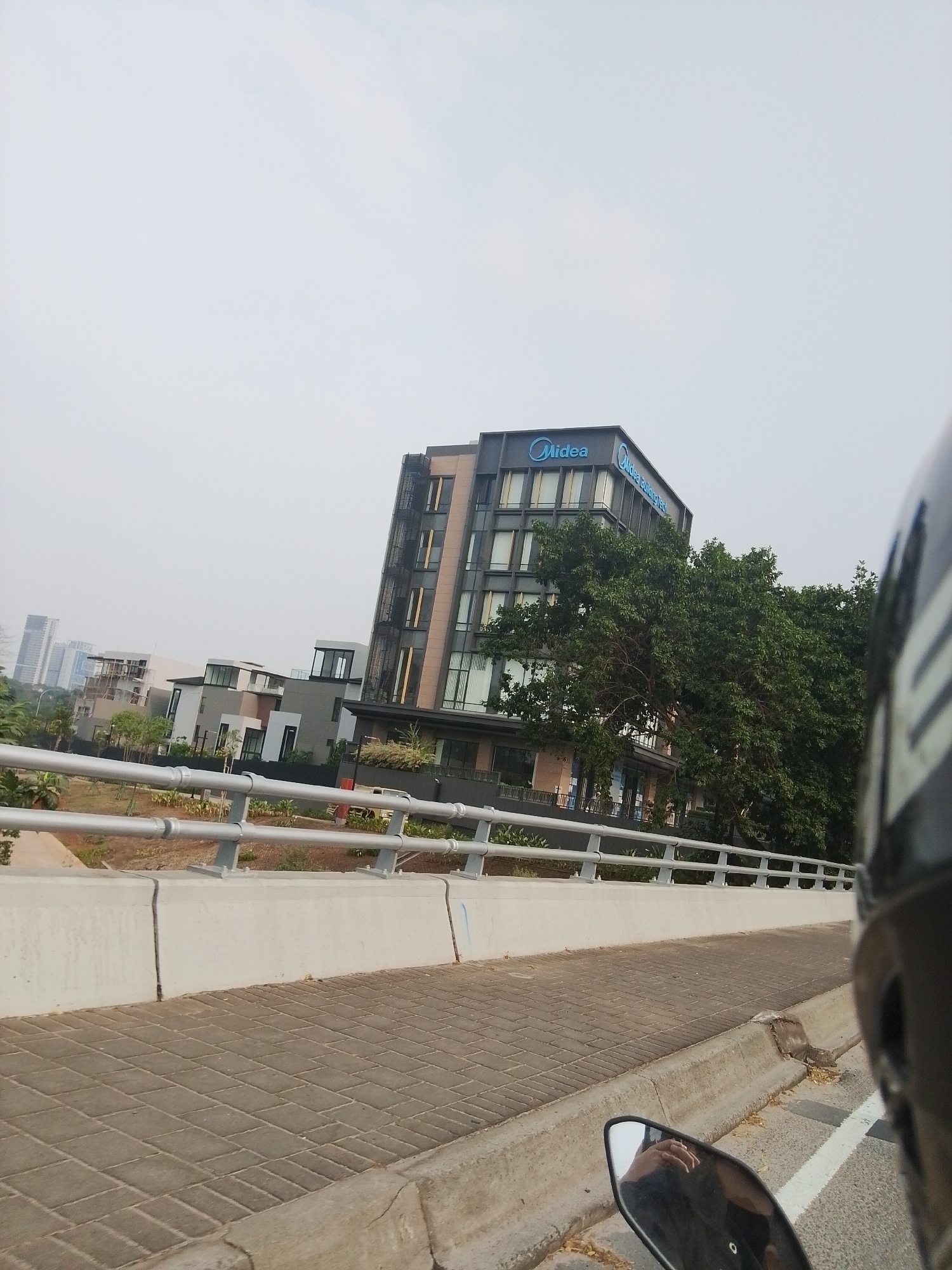 The image, captured from a motorcycle, shows a multi-story modern building with the "Midea" logo visible on its facade. The building is surrounded by greenery and appears to be located in a developed urban area. In the background, other buildings and a hazy sky are visible. The foreground of the image shows a paved walkway, a concrete barrier, and a metal railing. The reflection in the motorcycle's side mirror shows a person's hand. The location is identified as Lengkong Kulon, Indonesia.