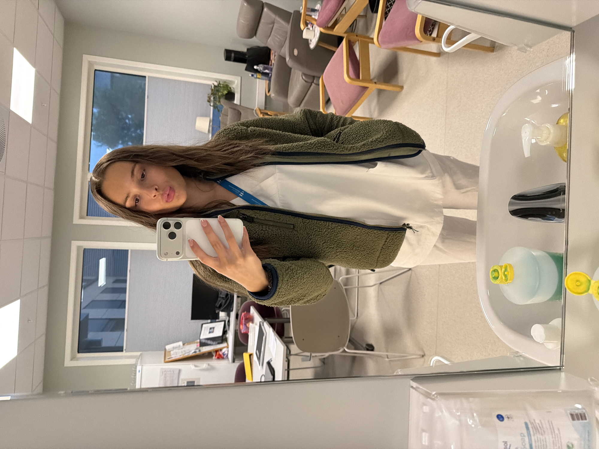 A young woman with brown hair is taking a mirror selfie in what appears to be a clinical or office environment. She is wearing a white V-neck scrub top and off-white scrub pants, with a green fleece jacket over the top. She is holding a white smartphone in her right hand, taking the picture. The background visible in the mirror shows a room with windows, office furniture including chairs and a desk, and medical equipment like recliner chairs. There is a sink in the foreground of the mirror reflection with soap dispensers and bottles. The scene is well-lit, suggesting an indoor setting. The location is identified as Harstad, Norway.