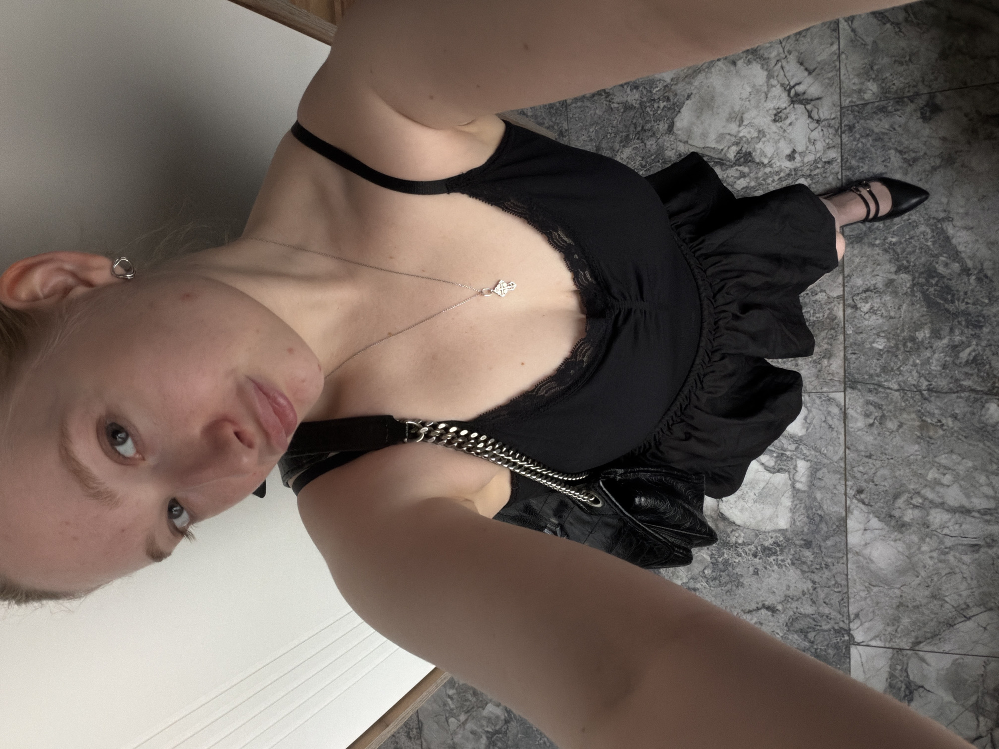 The image shows a woman wearing a black dress, black shoes, and a black purse. She has blonde hair and is wearing a necklace. The scene appears to be indoors, with marble flooring visible. The lighting suggests it could be daytime. The woman is looking at the camera with a neutral expression. The image is a selfie, taken from a high angle, looking down at the woman.