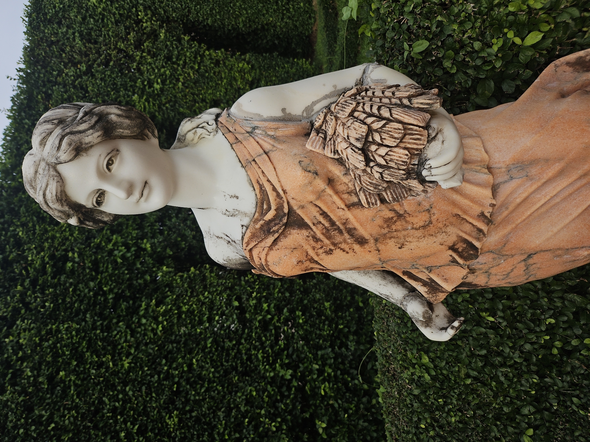 A white marble statue of a woman in a draped orange garment stands in front of a dense green hedge wall in Na Chom Thian, Thailand. The statue depicts a young woman with her hair styled in curls, her head tilted slightly to the left, and her gaze directed upward. She is holding a bundle of what appears to be wheat or grain in her right arm. The statue shows signs of weathering, with dirt and discoloration visible on its surface. The hedge wall behind the statue is meticulously trimmed and forms a lush green backdrop. The ground in front of the hedge is also covered in low-lying green foliage. The sky above is overcast, suggesting a cloudy day. The overall scene is one of serene, classical beauty set within a cultivated garden environment.