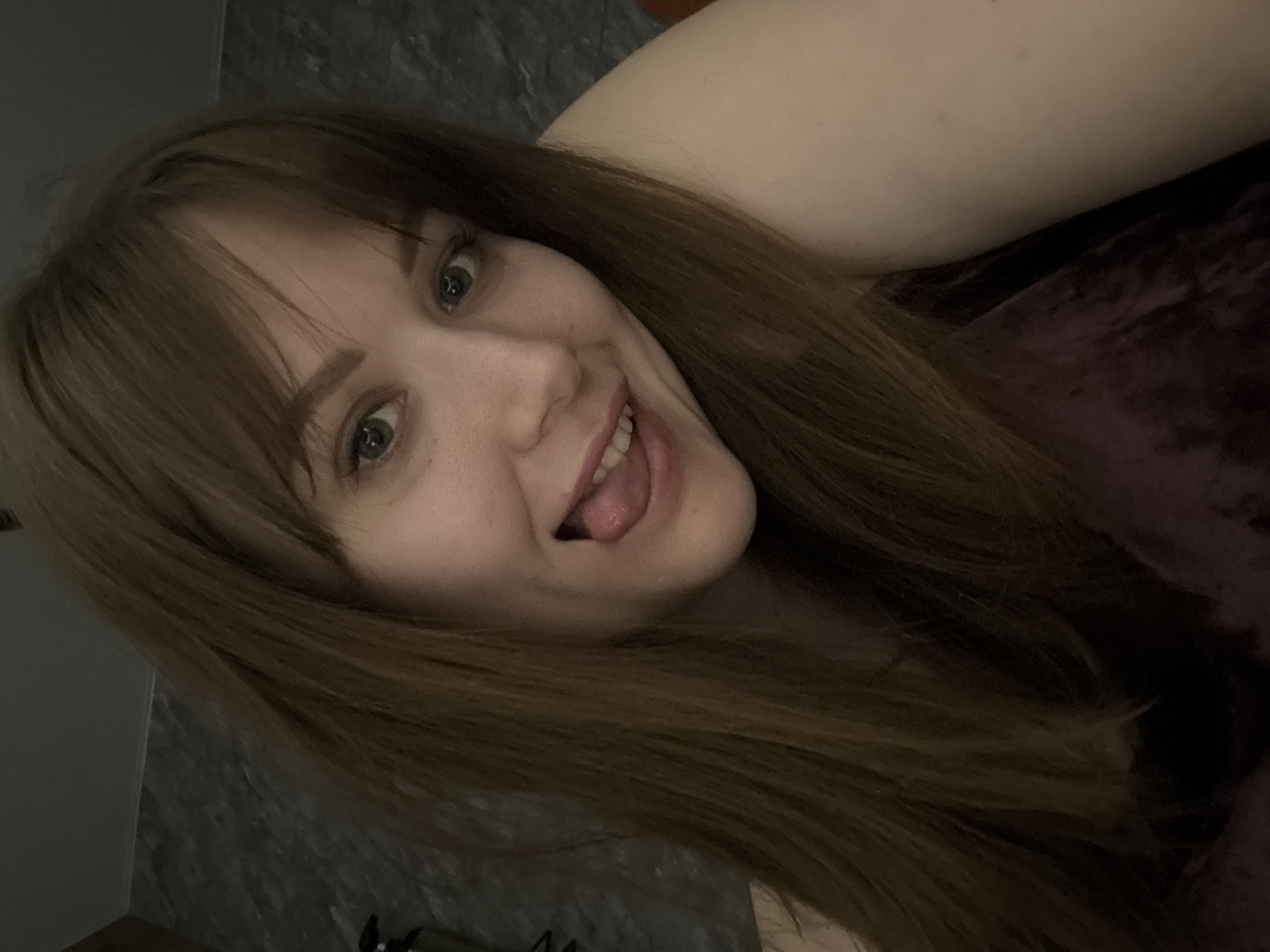 A woman with light skin and brown hair is posing for a selfie. She is lying down, her head tilted, and her tongue sticking out. She has green eyes and some freckles on her face. She is wearing a dark purple top. The background includes a grey textured wall and a glimpse of a lighter-colored surface. The location is not discernible from the image.