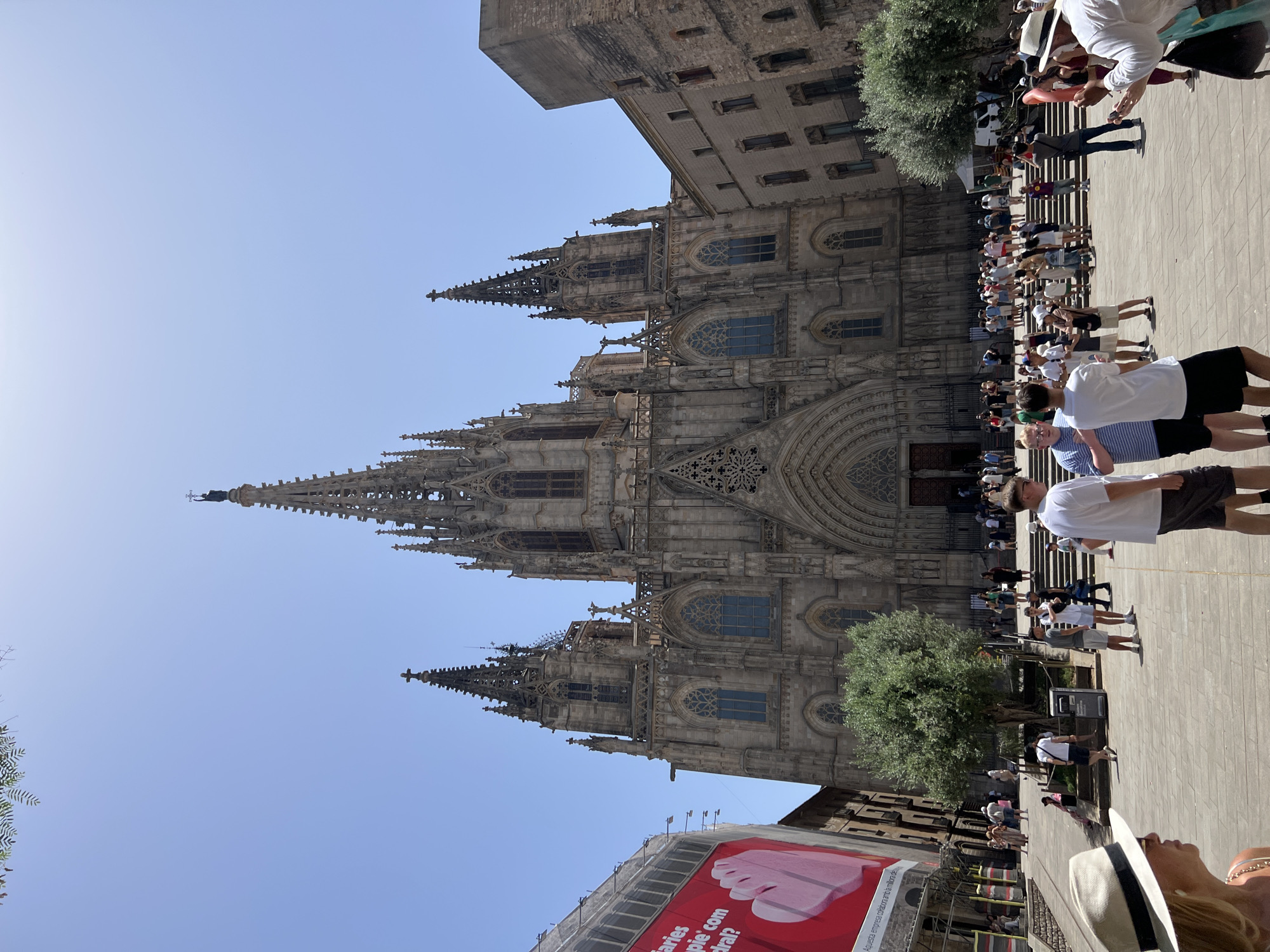 The image shows the Barcelona Cathedral in Barcelona, Spain, on a sunny day. The cathedral is a large Gothic structure with intricate stonework, spires, and arched entrances. In front of the cathedral, there is a large public square paved with stone tiles. Many people are present in the square, some walking, others standing in groups or sitting on steps. Some are looking towards the cathedral, while others are engaged in conversations. A large red billboard is visible on the left side of the image, with some text and a graphic of a hand. Trees are planted in the square, and there is some scaffolding on a building to the left. The sky is clear and blue.