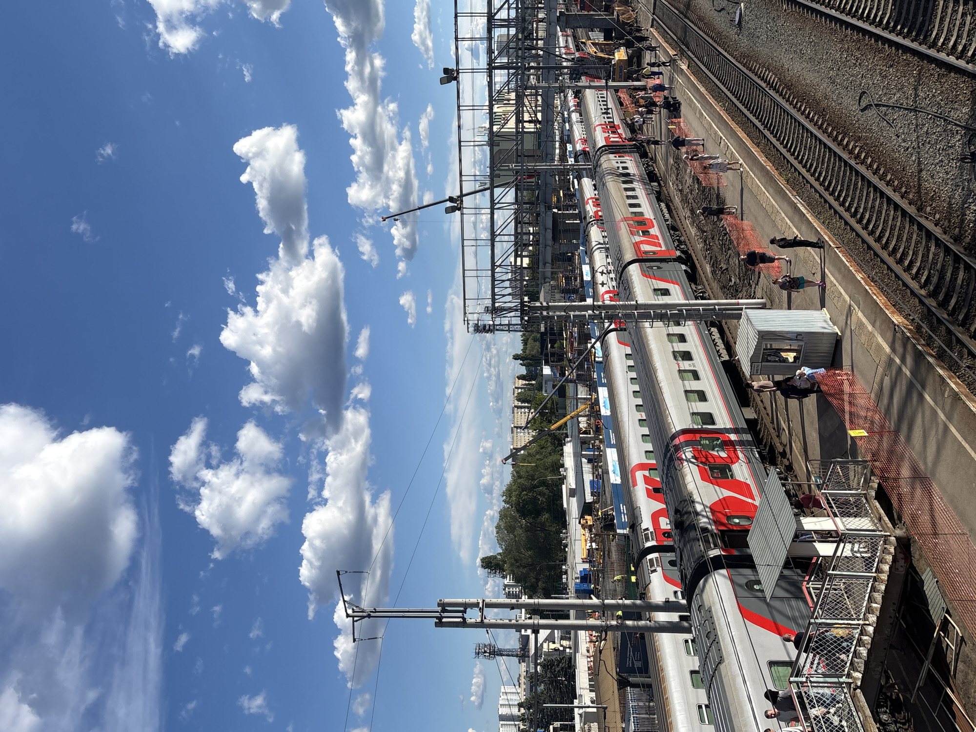 The image shows a train platform in Saratov, Russia on a sunny day with a bright blue sky and scattered white clouds. Three parallel train tracks are visible, with a passenger train occupying one of them. The train is white and grey with red accents and the Russian Railways logo. People are standing on the platform, some appearing to board or disembark the train. Construction activity, including cranes and equipment, can be seen in the background, suggesting ongoing development or maintenance work. The architecture in the background features a mix of mid-rise buildings and trees, typical of an urban setting.