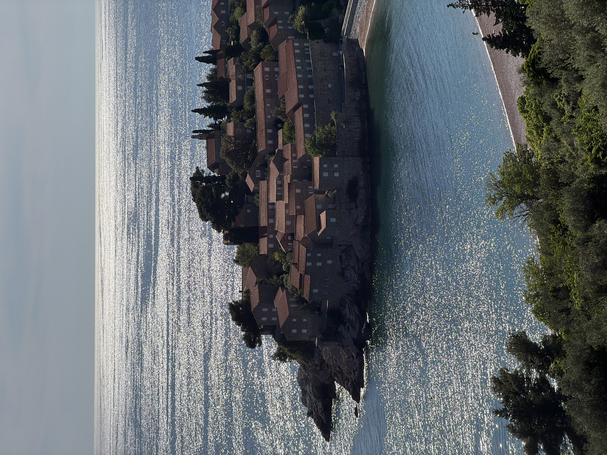 The image depicts a coastal view of Sveti Stefan, Montenegro. The foreground is dominated by a peninsula covered in stone buildings with red-tiled roofs and interspersed with trees. The structures appear clustered together, forming a compact and historic settlement.
The peninsula is surrounded by the Adriatic Sea, which reflects sunlight, creating a shimmering effect across the water's surface. A beach is visible to the right of the peninsula. Vegetation is seen on the edge of this beach.
The sky is bright and partially clouded. The image is taken from an elevated position, offering a panoramic perspective of the landscape.