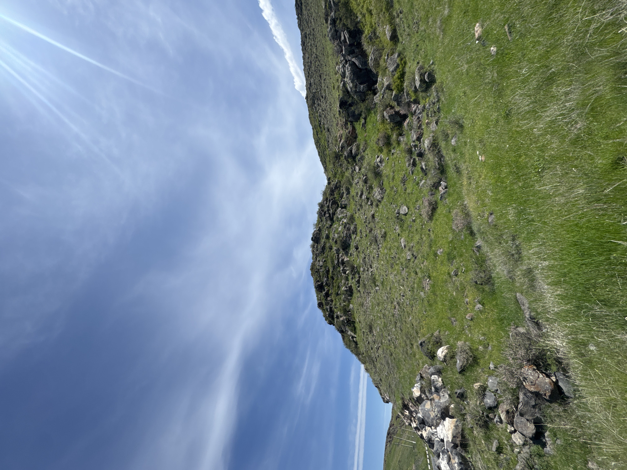 The image shows a hillside in Bagravan, Armenia. The hillside is covered in green grass and scattered rocks. The sky is blue with streaks of white clouds. The sun is shining brightly, creating a lens flare effect in the upper left corner of the image. The camera is oriented at an unusual angle, giving the impression that the hillside is tilted. In the lower portion of the image there is a fence.