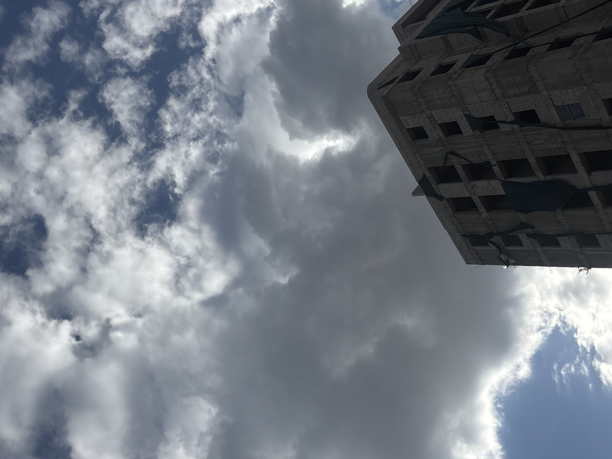 The image is an upward-angled view featuring a partially constructed building against a backdrop of cloudy skies. The building, located in Yerevan, Armenia, is in a raw concrete state with visible rectangular window openings, some covered with black netting. The sky is a mix of blue and grey, with prominent cloud formations. The composition is oriented with the building occupying the upper right quadrant of the image, while the sky and clouds fill the remaining space. The environment suggests an urban setting under overcast weather conditions.
