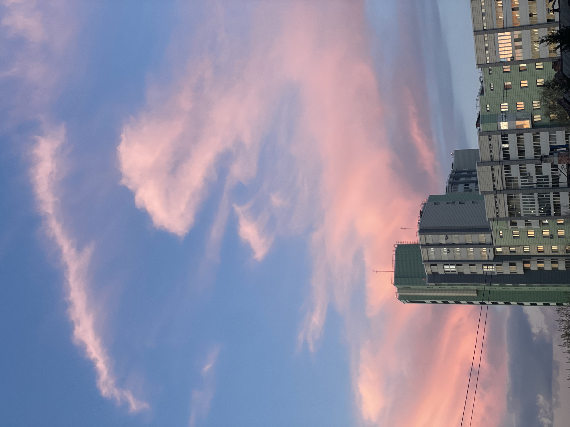 The image shows a view looking up at several tall buildings against a sky with pink-tinged clouds. The buildings are a mix of grey and green and have multiple windows, some of which are lit. The sky is blue with pink and white clouds. The image is rotated 90 degrees, so the buildings are oriented vertically on the right side of the frame. It is unknown if this is a building located in the city of Krasnoyarsk.