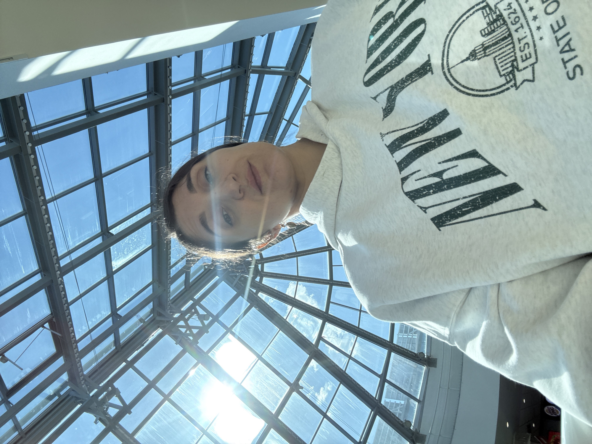 The image shows a person looking up towards a glass ceiling with a metal framework. The sunlight shines through the glass panes. The person is wearing a light-colored sweatshirt with a "NEW YORK STATE OF" logo and EST. 1624. The logo includes an image of the Empire State Building. The location appears to be an enclosed atrium or public space with a glass roof. I cannot confirm the location in the city of Tobolsk, Russia.