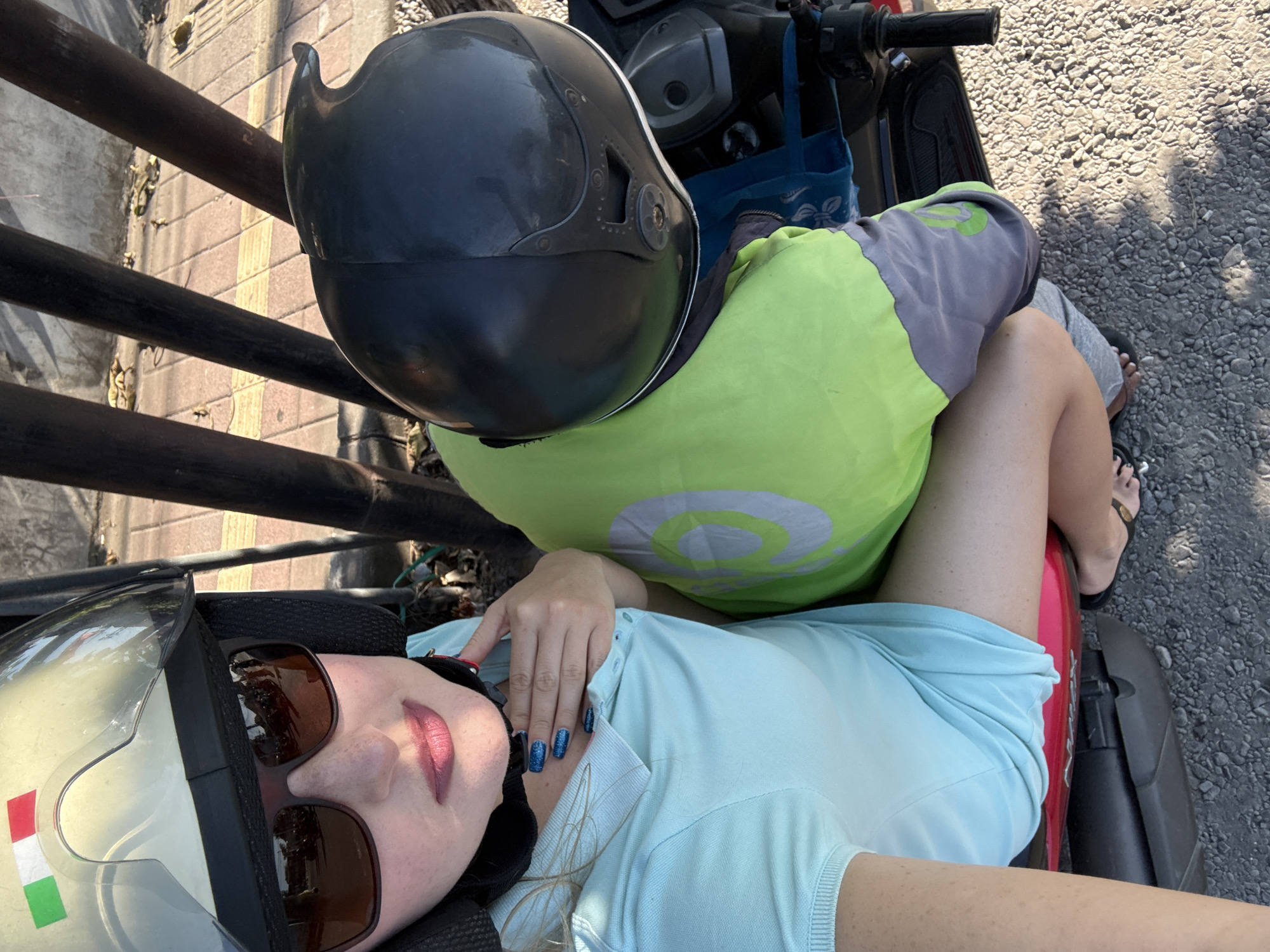 A woman is seated on a scooter in Denpasar, Indonesia, with her left hand resting on her chest and her right hand holding onto a green pillow. She wears sunglasses and a light blue polo shirt, with her fingernails painted blue. A white helmet with an Italian flag sticker is on her head, and a black helmet is beside her. The scooter's red seat is visible beneath her legs, and she is wearing black flip-flops. The ground is covered in small gray pebbles, and a sidewalk with patterned tiles is visible in the background. Several dark, vertical poles are also present.