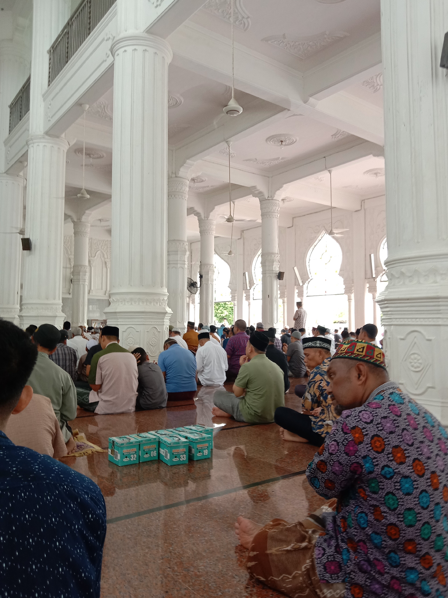 A wide-angle, medium shot shows the interior of a mosque in Lhokseumawe, Indonesia, with a group of Muslim men seated on the floor for prayers. The men, of varying ages, wear traditional clothing, including skullcaps and batik shirts. They are arranged in rows facing away from the camera.
Several rectangular boxes, possibly containing prayer beads or other religious items, are positioned on the floor in the foreground. The mosque's interior is characterized by white walls, tall Corinthian-style columns, and decorative moldings on the ceiling. Ceiling fans are visible. Light floods the room through arched windows. The floor is made of reddish-brown tiles.
